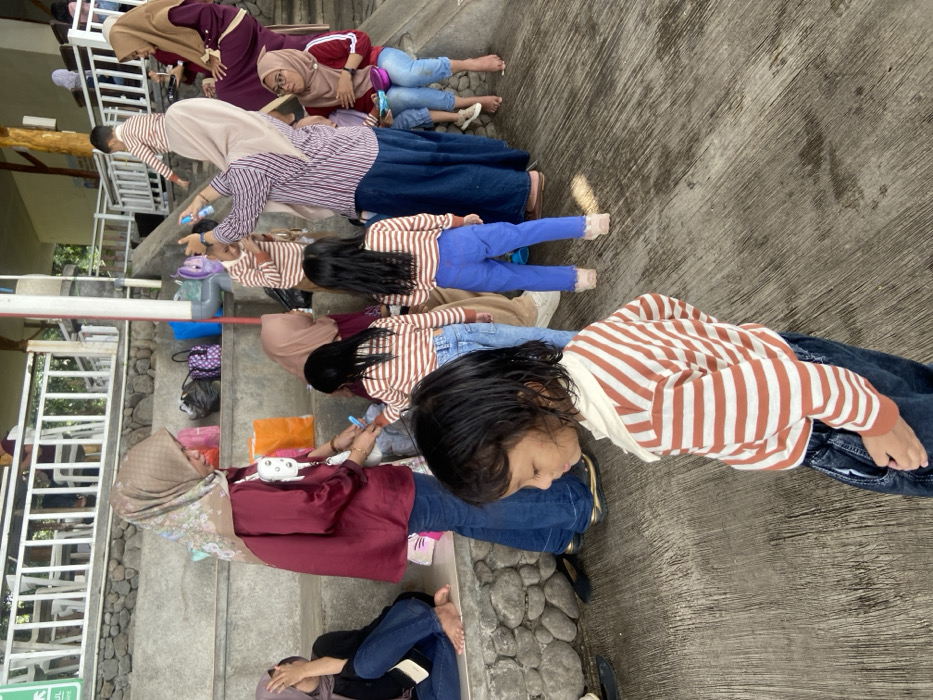 A group of people, mostly women and children, are lined up in a row on steps and a concrete area. Many of the women wear hijabs. Some are bent over looking at their hands or feet. The children are mostly wearing striped shirts and blue pants or jeans. They are barefoot and facing the same direction. The scene suggests an outdoor activity or gathering.
