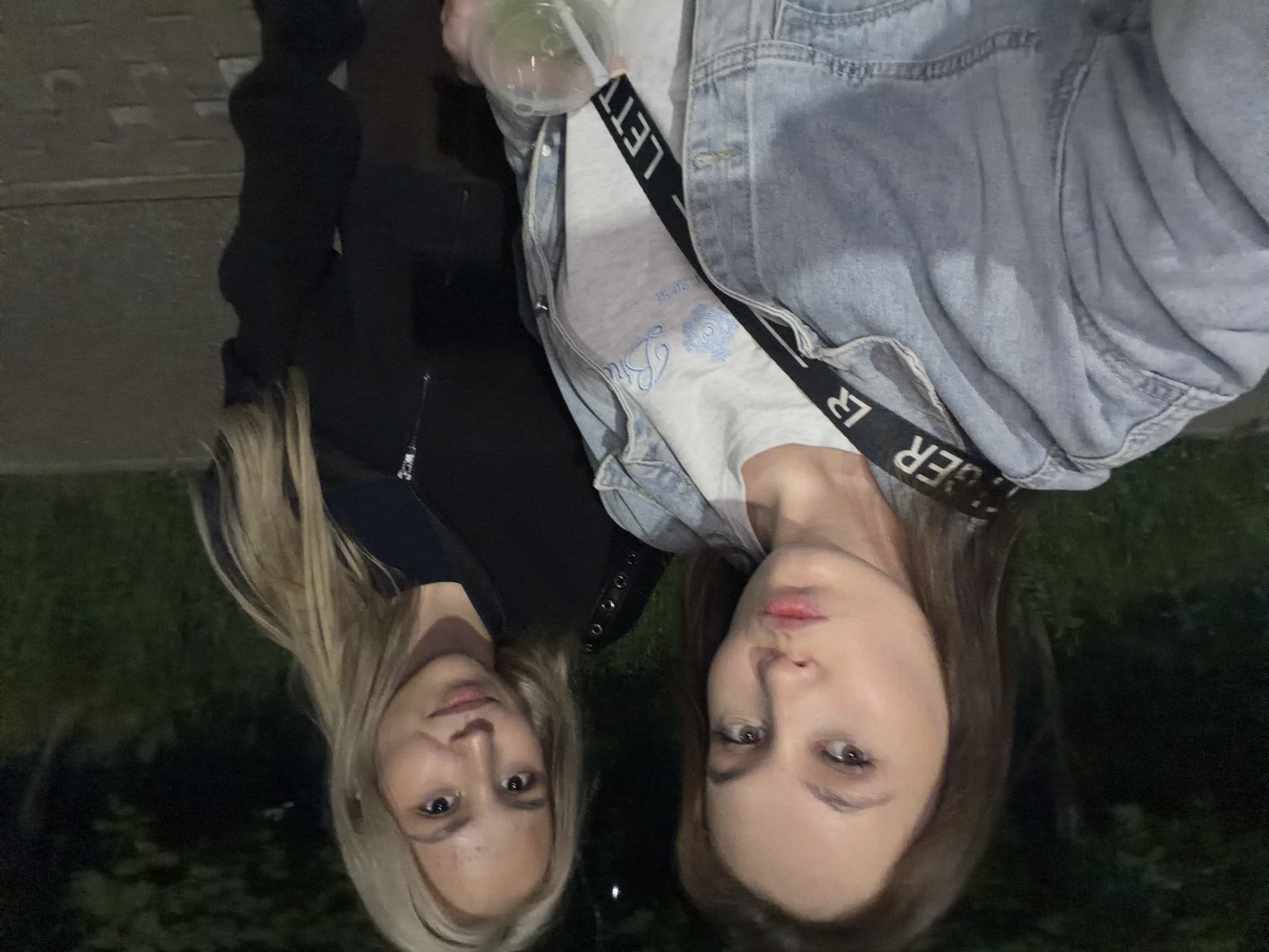 Two young women are photographed outdoors at night in Naberezhnye Chelny, Russia. The woman in the foreground, on the left, has long brown hair and is wearing a light blue denim jacket over a light-colored t-shirt with blue embroidery. A black strap with white lettering crosses her chest. She is puckering her lips. The woman on the right has long blonde hair and is wearing a black zip-up jacket. She is looking towards the camera with a neutral expression. They appear to be standing on a paved surface with some greenery in the background. The lighting suggests an urban environment at night.