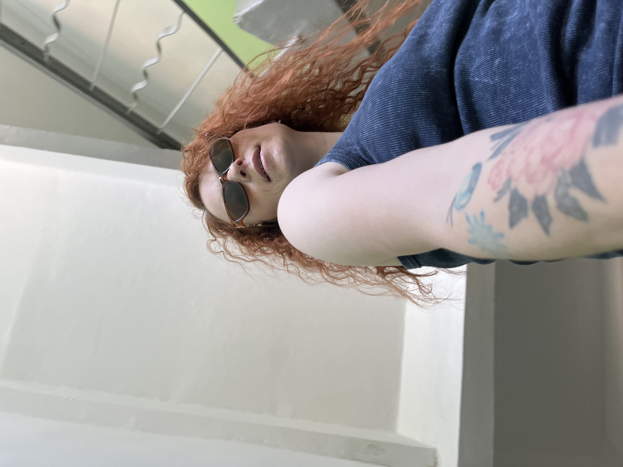 A person with reddish-brown curly hair, wearing sunglasses and a dark blue top, poses for a close-up, slightly tilted photograph. The person has tattoos on their left arm. The background consists of a white wall and a railing. The image was possibly taken in the city of Chaikovsky, Russia.