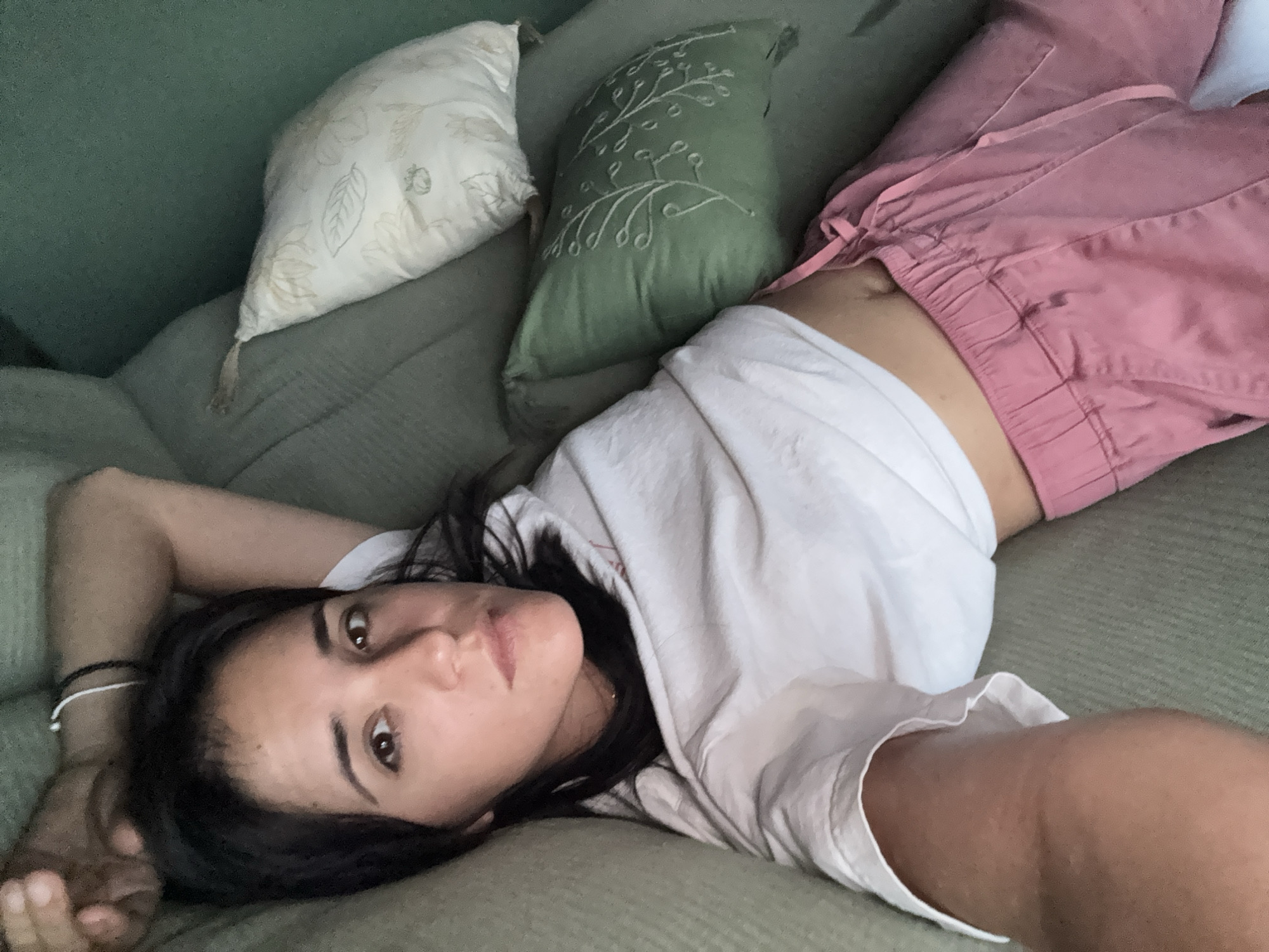 A young adult woman is lying on a green couch. She is wearing a white t-shirt and pink pants. Two pillows are visible behind her. The image was likely taken indoors. The location in the city of Huixquilucan de Degollado, Mexico cannot be determined from the image.