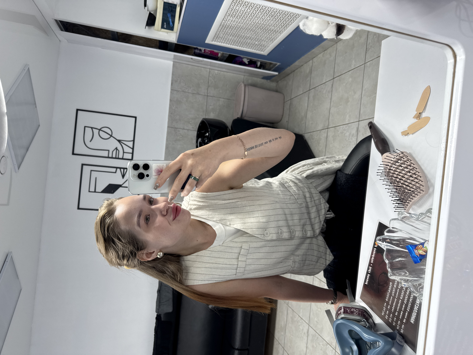 A woman is taking a selfie in a mirror, likely in a hair salon setting. The mirror reflects a portion of the salon, which appears to have white walls, black furniture, and framed artwork. On the table next to the mirror, there are items such as a hairbrush, hair clips, a dish with candy, and printed materials, possibly advertisements or informational pamphlets. The woman is holding a smartphone with multiple camera lenses. Her attire includes a striped top and visible jewelry like a ring and bracelet. The OCR text appears to contain Russian language text from the printed materials, suggesting the location could be Chaikovsky, Russia.