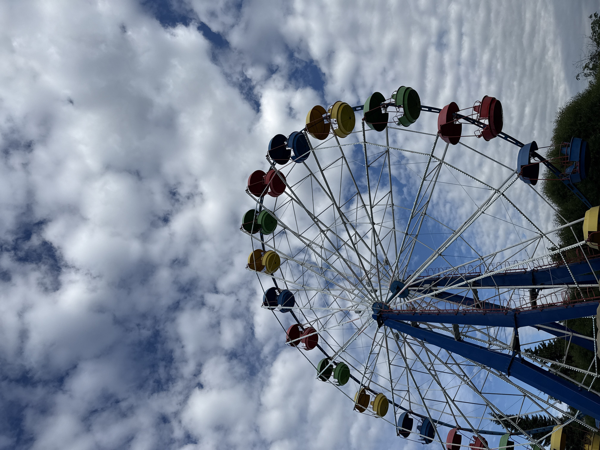 The image shows a Ferris wheel in an outdoor setting. The Ferris wheel has a blue central support structure and is adorned with colorful gondolas, including red, yellow, blue, and green. The wheel is seen against a backdrop of a cloudy sky, with patches of blue visible. In the lower part of the image, glimpses of green trees and foliage suggest a park or amusement area. The overall scene is that of a recreational attraction under a partially cloudy sky. No specific location in Saint Petersburg, Russia is identifiable from the image itself. There are no people or discernible actions in the image.