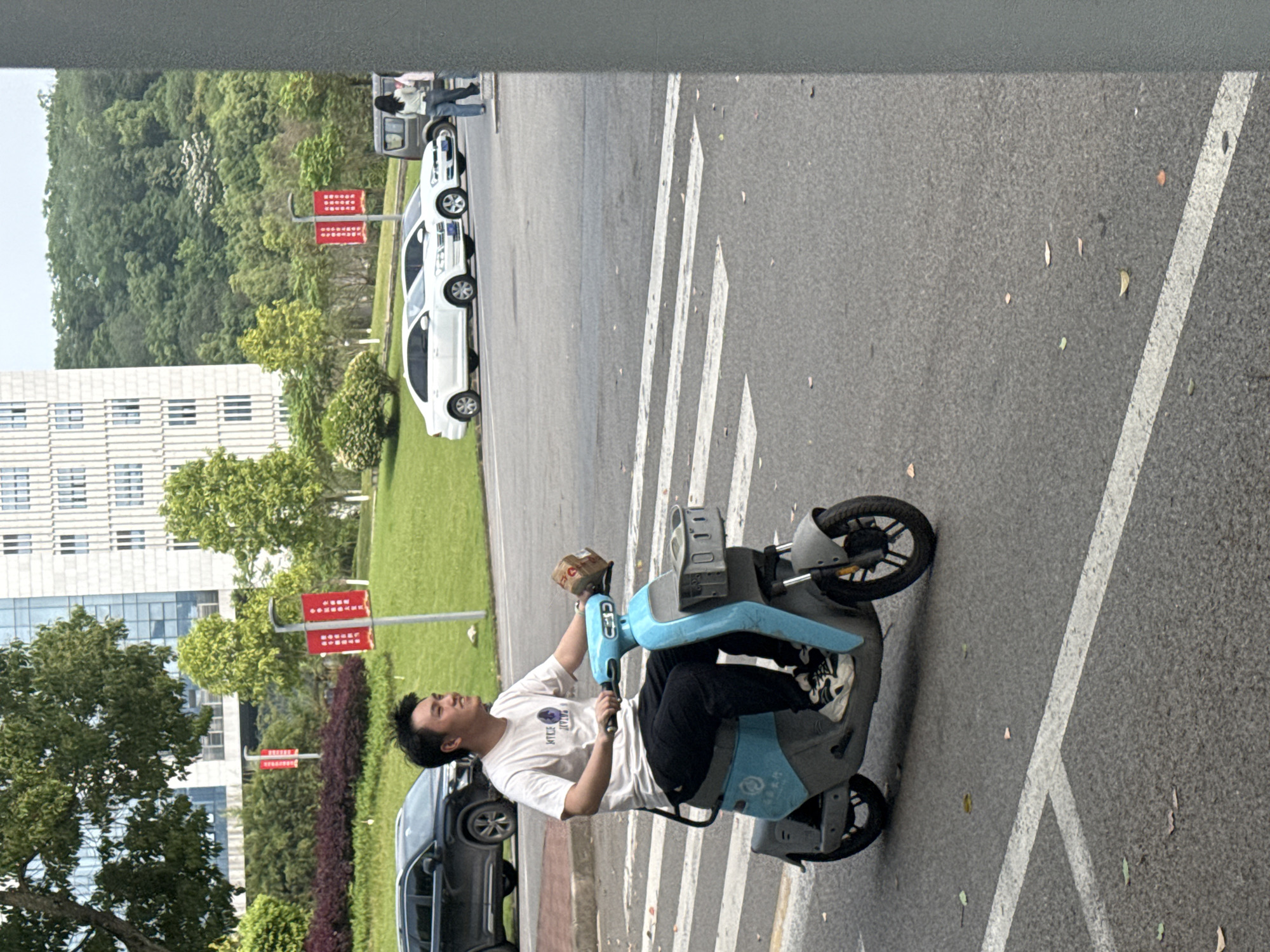 A man is riding a light blue electric scooter on an asphalt road. He is wearing a white t-shirt and black pants and is holding a small cardboard box. The road has white painted lines marking crosswalks or lanes. The background shows trees, a building, parked cars, and red signposts. The location appears to be in Jiangjin, China.