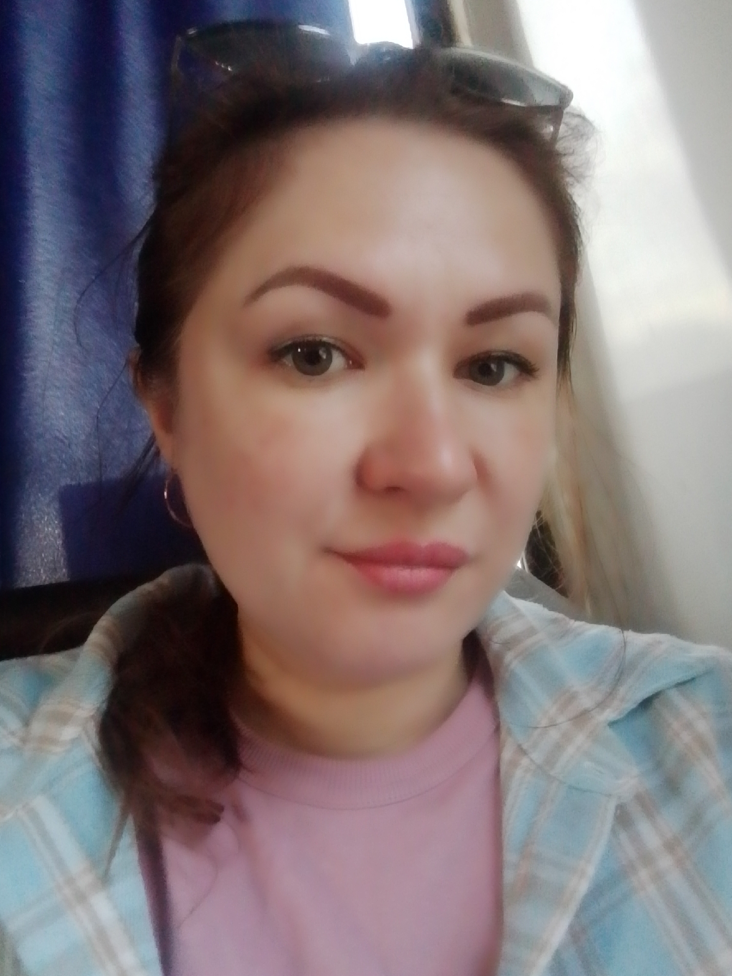 A head-and-shoulders shot captures a woman with brown hair partially pulled back. She has sunglasses perched on her head. She's wearing a pink shirt with a light blue plaid top. Her skin is fair, with reddish undertones visible on her cheeks. She is wearing light makeup, including eyebrow shaping, eye shadow, and lip color. The background is divided with a blue drape or curtain to the left and a pale, neutral wall to the right. There is no indication of the location in the city of Chaikovsky, Russia.