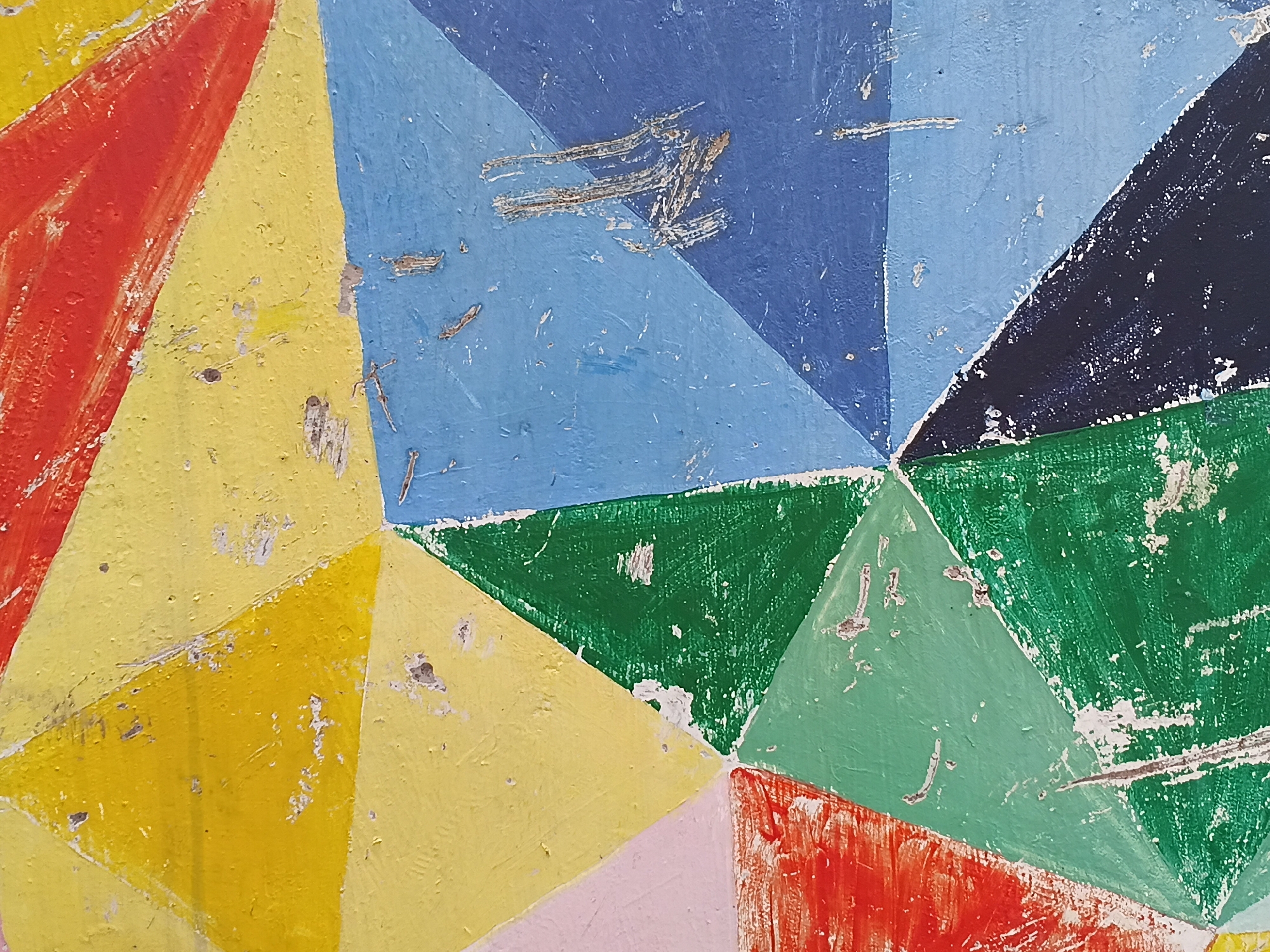 This is a close-up, abstract photograph depicting a wall or surface adorned with a geometric pattern of painted triangles, located in Shunde District, China. The pattern is composed of numerous triangular segments in a vibrant palette, including bright red, yellow, various shades of blue (light and dark), green, light green, and subtle hints of pink.
The paint shows significant signs of wear and age, characterized by extensive chipping, flaking, and deep scratches that expose the lighter, textured substrate beneath. Visible brush strokes contribute to the rough, uneven surface texture. The triangles are arranged in a tessellating mosaic, with several converging at central points, creating a fragmented and dynamic visual effect. There is no visible activity, event, or text in the image. The distressed state of the paint suggests prolonged exposure to elements or heavy use, highlighting a worn and weathered aesthetic.