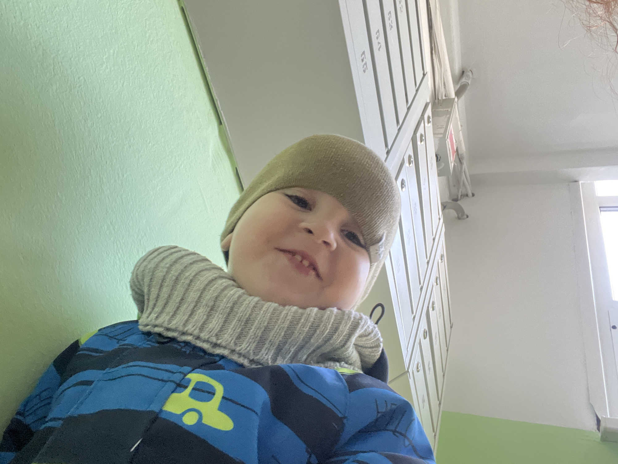 A young child wearing a tan knit hat, a striped gray neck gaiter, and a blue and black striped jacket with a yellow car design is looking up at the camera. They appear to be indoors, standing next to a pale green wall. To the right, there's a row of white mailboxes with numbers on them, ascending vertically. The background also shows a white ceiling and a window. The location is the city of Chaikovsky, Russia.