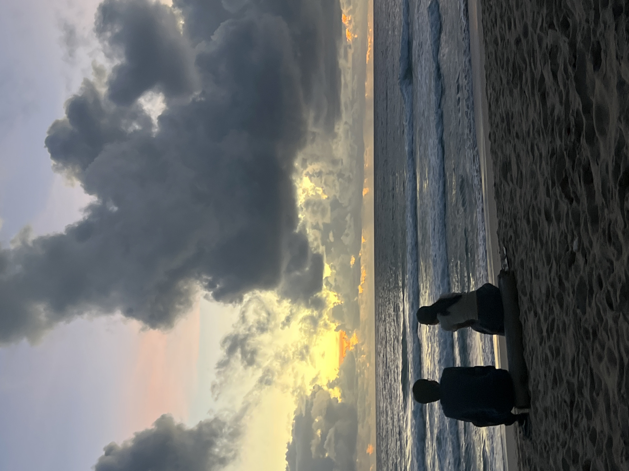 Two individuals, one identified as male and the other as female, are seated on a log on a sandy beach, facing the ocean. The male, on the left, wears a dark long-sleeved shirt and dark pants. The female, on the right, is in a light-colored short-sleeved top and dark pants. They are positioned in silhouette against a dramatic sky at dusk or dawn. The ocean exhibits moderate waves and the sky is filled with large, dark storm clouds interspersed with patches of soft yellow and pink light, suggesting either sunrise or sunset. The sand in the foreground is dark and textured, with footprints visible. The overall scene depicts a tranquil moment of observation of the natural elements. The location is identified as Choeng Thale, Thailand.