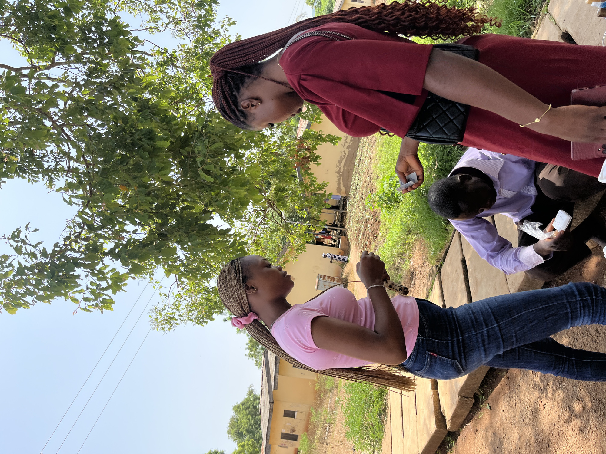 Two young women with braided hair are standing and interacting outdoors in Rafin Par, Nigeria. One woman on the left wears a pink t-shirt and blue jeans. The other woman on the right wears a maroon dress and a black quilted handbag. Both women are looking down at something they are holding in their hands. In the foreground, a man wearing a purple collared shirt is kneeling and looking down at an object. The background shows a yellow building with a tiled roof, green vegetation, and a large tree. The ground is composed of dirt and some paving stones. The sky is clear and blue.