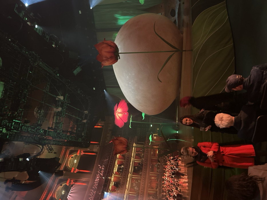 The scene depicts a theatrical performance with an elaborately decorated stage. Oversized flowers and a large egg-shaped object are visible. Seated audience members watch the show. A woman in a red coat is noticeable in the foreground. Complex stage rigging and lighting contribute to the spectacle. The overall atmosphere suggests a whimsical or fantastical theme.