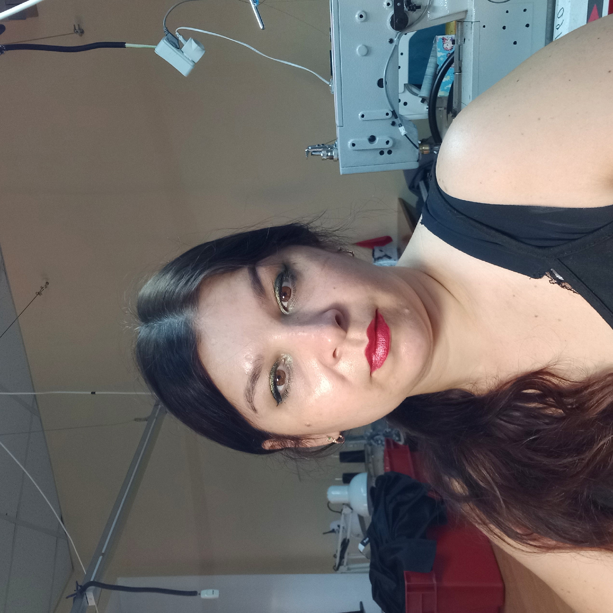 A medium shot captures a woman with long, dark, wavy hair, wearing a black tank top and red lipstick, posing for a selfie in what appears to be a workshop or studio. She is looking directly at the camera with a neutral expression. Her makeup includes green and gold eyeshadow. In the background, to her right, is a white industrial sewing machine. To her left, there is a red plastic bin filled with dark fabric. Overhead, fluorescent lighting fixtures and electrical cords are visible on the ceiling. The walls are a neutral, light brown color. The overall scene suggests a workspace, potentially for sewing or garment production, located in the city of Kropyvnytskyi, Ukraine.