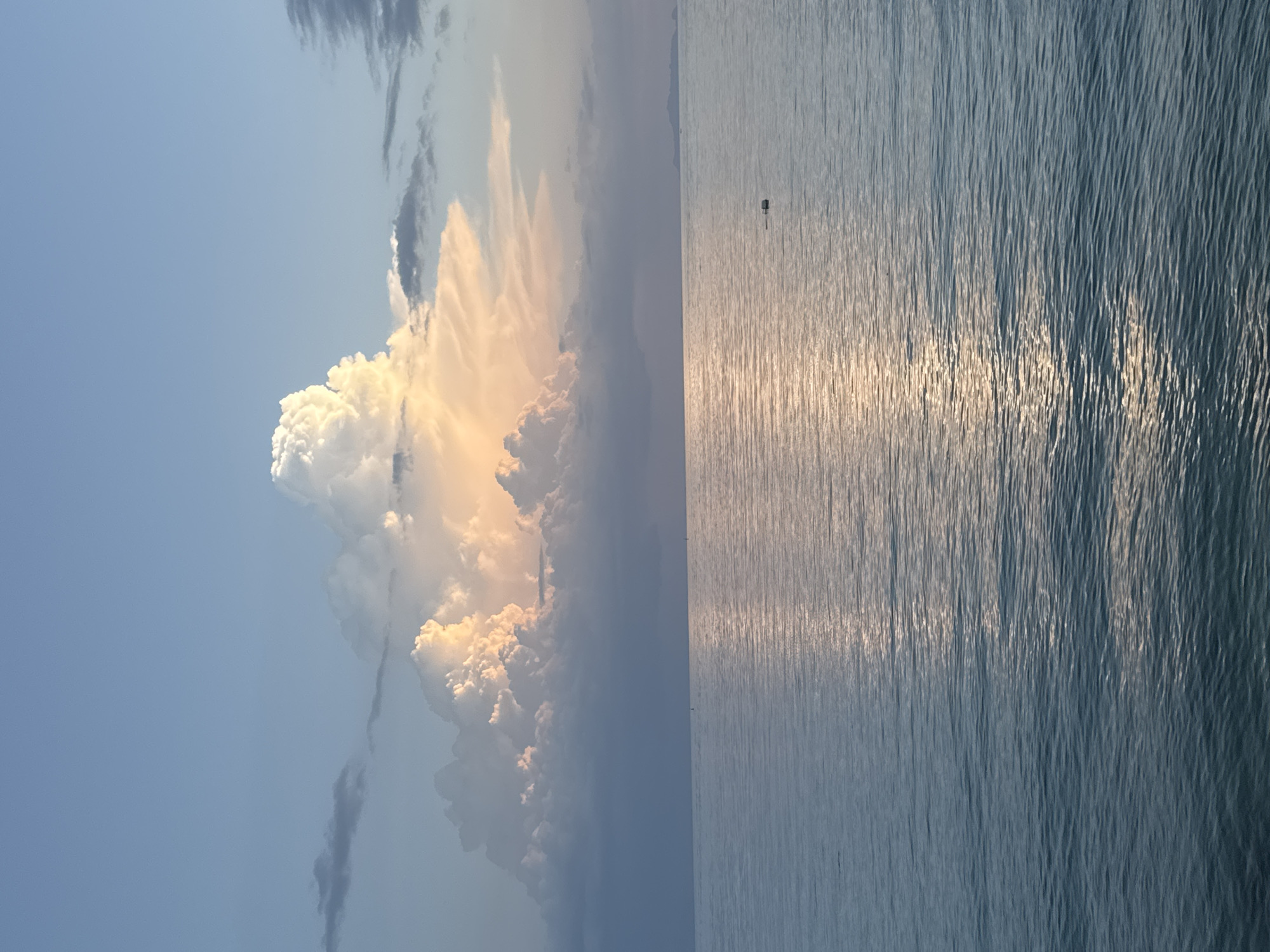 A serene seascape featuring a vast expanse of calm, light blue ocean under a sky of pale blue. Dominating the upper portion of the image is a massive, illuminated cumulonimbus cloud formation. The cloud is a dramatic spectacle of light and shadow, with its upper sections glowing brilliantly white and a warm, peachy-orange hue, suggesting it is either catching the last rays of a setting sun or the first light of dawn. Darker, more ominous clouds gather below, creating a stark contrast. The sun's reflection shimmers across the water's surface, creating a bright, silvery path. A small, dark marker buoy is visible in the middle of the ocean. In the far distance, a faint landmass or island is visible on the horizon. The scene evokes a sense of tranquility and the majestic power of nature. This scene likely depicts the coast of Ko Pha-ngan District, Thailand, known for its beautiful beaches and dramatic sunsets.