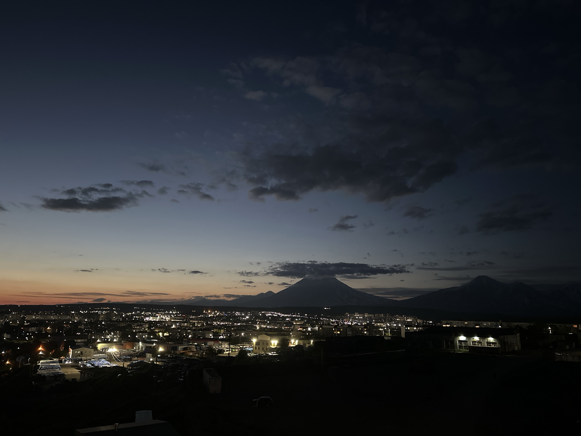 The image shows a panoramic view of the city of Petropavlovsk-Kamchatsky, Russia, at dusk. The sky transitions from a deep blue at the top to a soft orange and pink near the horizon, with scattered clouds. The city lights are illuminated, creating a vibrant glow against the darkening landscape. In the background, the majestic volcanoes of Avachinsky and Koryaksky stand silhouetted against the twilight sky, with a few clouds clinging to their peaks. The foreground is dark, obscuring details of the terrain and any potential inhabitants or activities. The scene is a serene and picturesque depiction of the city at the transition from day to night.