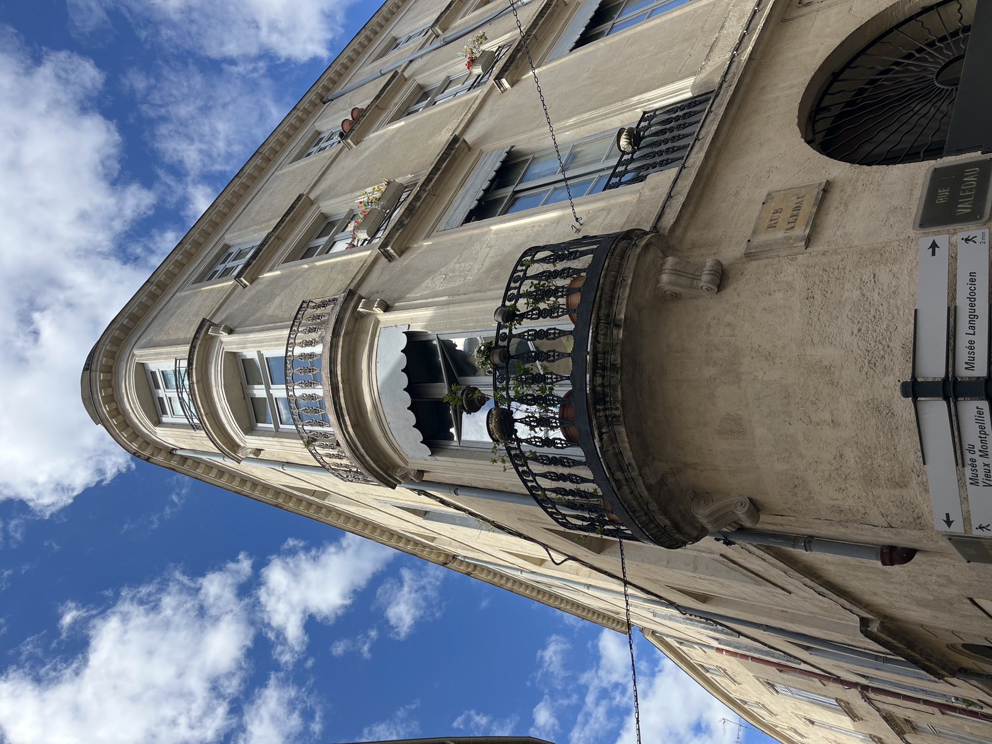 Looking upwards, the scene shows a pale-colored building exterior under a blue sky scattered with white clouds. The building has multiple windows with dark wrought-iron balconies, some adorned with flower boxes and plants. Signage on the building indicates "Rue Valedau" and directs toward the "Musée du Vieux Montpellier," confirming the location as Montpellier, France. There are no people visible in the frame. The building's architecture features curved corners and decorative elements. A drainpipe runs vertically down the building facade.