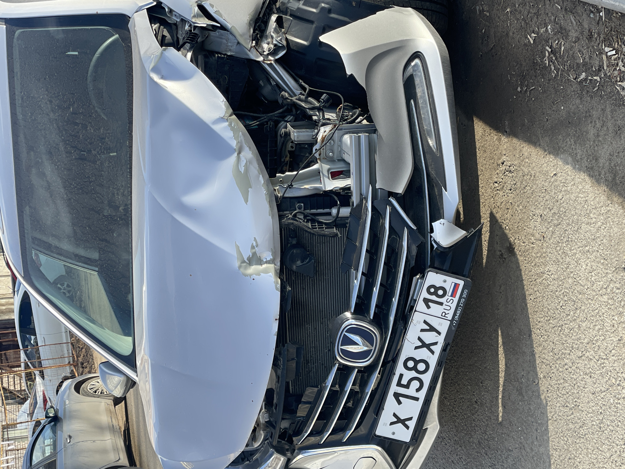 The image displays a white car that has sustained severe front-end damage, indicative of a significant collision. The car's hood is heavily crumpled and pushed upwards, exposing the internal components of the engine bay. Wires, structural elements, and the radiator are visible through the mangled front end. The front bumper is largely detached and fractured, with parts missing or hanging, and the grille is also broken, though the central car emblem, appearing to be a Changan logo, remains relatively intact.
A Russian license plate is attached to the damaged front, clearly displaying the characters "X 158 XY 18 RUS". Below the "RUS" marking, a phone number "+7 (8482) 270 300" is also present. The "18" in the license plate signifies the Udmurt Republic, confirming the location context of Izhevsk, Russia.
The scene is set outdoors under bright daylight, as evidenced by strong sunlight and distinct shadows cast on the paved ground. The ground surface appears to be asphalt or concrete, with some scattered debris like dirt and dry leaves. The car's windshield is visibly dusty. No individuals are present in the photograph. The visible damage suggests the vehicle is currently out of commission.