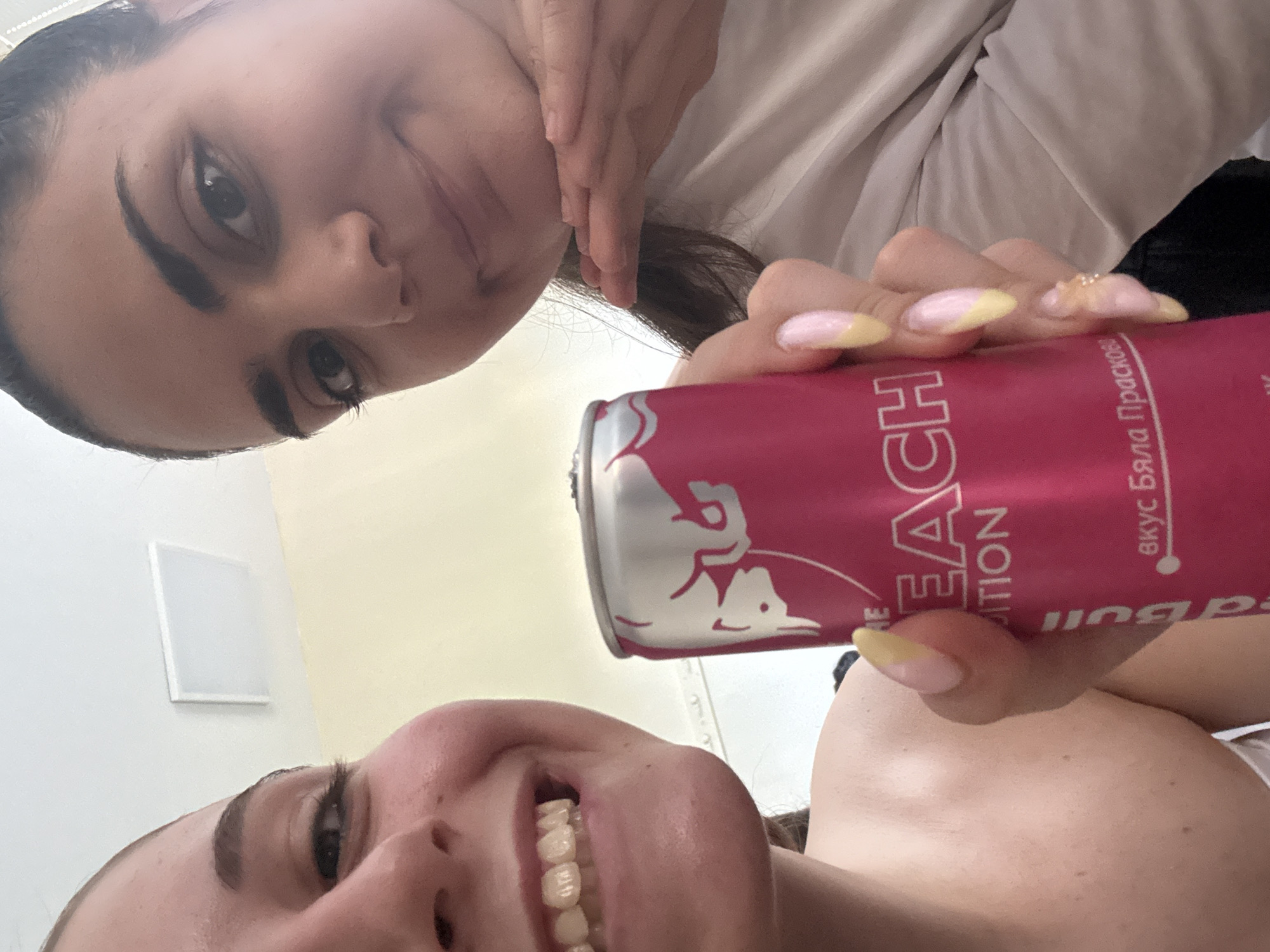 Two young women are in close proximity, possibly taking a selfie together. One woman is positioned above and to the left, while the other is below. Between them, a pink Red Bull can labeled "Peach Edition" in English and with text in a Cyrillic alphabet (possibly Russian) is visible. The location appears to be indoors, with a white wall and a square white electrical outlet visible in the background. There is no specific information that confirms this image was taken in the city of Burgas, Bulgaria.