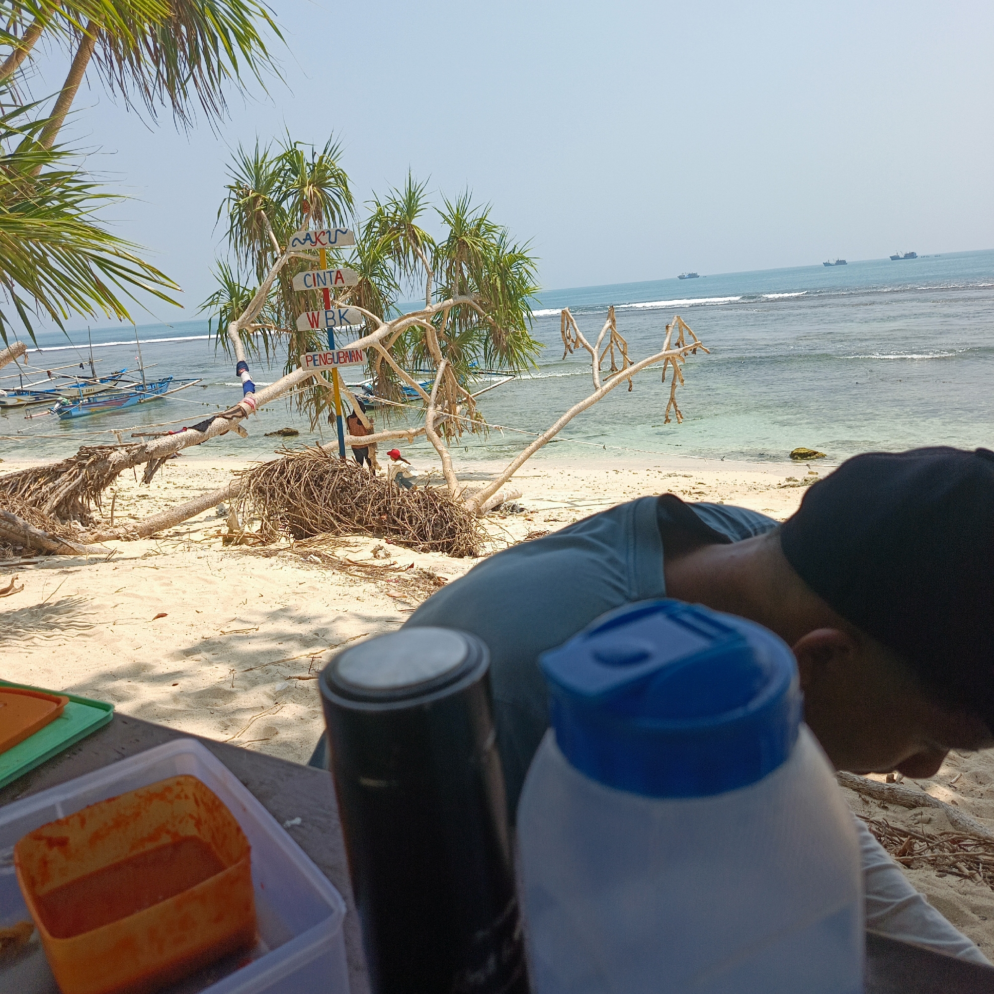 The image depicts a beach scene in Pengubaian, Indonesia. In the foreground, a person wearing a blue shirt and a black cap is partially visible, their head bowed. In front of them are various objects on a table, including a black cylindrical container with a silver top and a white bottle with a blue cap, likely containing liquids. An orange container with red contents sits to the left. The background reveals a sandy beach with scattered debris, including dried branches and roots from fallen trees. Beyond the beach, the ocean is visible with several boats anchored and a few larger vessels on the horizon. Palm trees and pandanus plants with spiky leaves are present on the beach. A directional signpost with multiple signs pointing in different directions, including one reading "CINTA" and another "PENGUAIAN," is positioned among the vegetation. The sky is clear and bright, indicating a sunny day. Two individuals, one wearing a red cap, are visible in the middle ground on the beach, near the signpost.