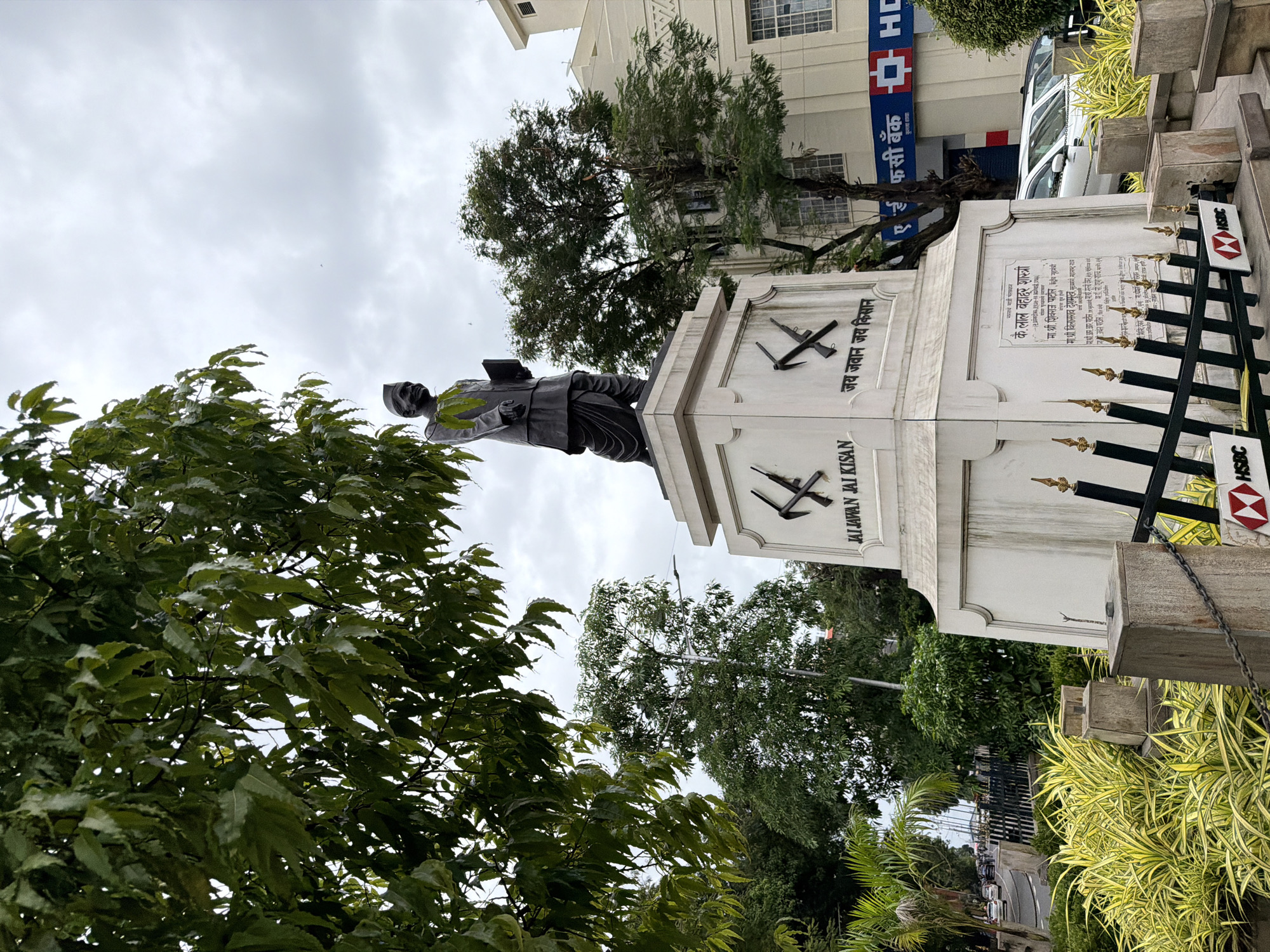 The image depicts a statue of a man in traditional Indian attire, likely a politician or significant historical figure, standing on a pedestal. The pedestal has inscriptions and symbols, including what appears to be agricultural tools and text in Hindi. The statue is located in an outdoor urban setting, with trees and foliage surrounding it. In the background, a building with the HSBC bank logo is visible, suggesting the location is in a commercial area. The sky is overcast, indicating a cloudy day. There are cars and a street visible in the distance, suggesting a public space within a city. The overall impression is a monument or memorial in an Indian city, possibly Mumbai, given the presence of HSBC bank and the style of the statue and surrounding architecture. The lower part of the image shows a black metal fence with a chain and decorative elements, along with more greenery and some concrete structures.