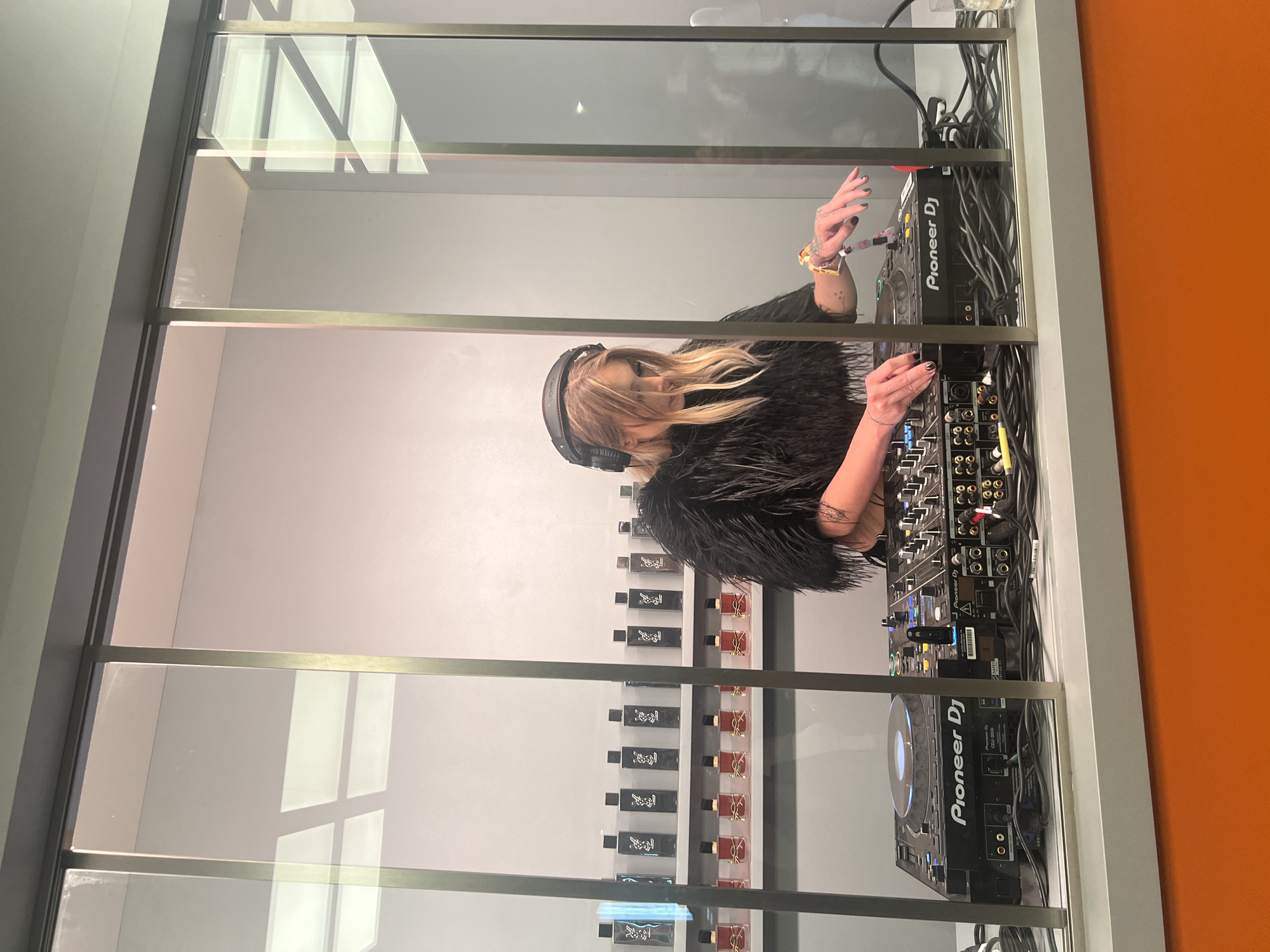 A DJ with headphones, wearing a black feathered jacket, is seen through a glass partition with metal framing. She is operating a Pioneer DJ setup consisting of a mixer and two CDJ units, all connected with numerous black cables. The backdrop includes a white wall with shelves displaying rows of perfume bottles and an orange wall to the right. The scene is located within a retail store in Indio, California, United States of America.