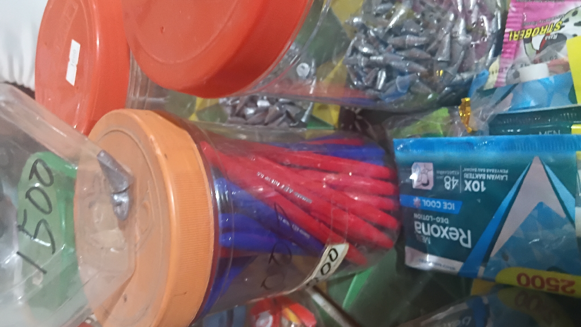 The image shows a close-up of several clear plastic containers filled with various items. In the foreground, a package of Rexona Deo-Lotion "Ice Cool" is visible, featuring a blue and white design with the text "10x LAWAN BAKTERI 48 PENYEGAR BAU BADAN". To the left of this package, another yellow package with "2500" is partially visible.
Behind the Rexona package, a clear plastic container is filled with red and blue pens. The lid of this container is orange, and a small metallic object, possibly a fishing sinker, is resting on top of the lid, secured by a clear plastic bag. A handwritten label on the bag appears to say "1500".
To the right of the container with pens, another clear plastic container holds a large quantity of small metallic objects, which resemble fishing sinkers or weights. This container also has an orange lid. Further to the right, another container with an orange lid is visible, but its contents are less clear.
The background is blurred but suggests a tabletop surface with some green and yellow patterns. The overall scene appears to be an indoor setting, possibly a shop or storage area. There are no people visible in the image.
The information about the specific location in Prabumulih, Indonesia, cannot be determined from the visual content of the image alone.
