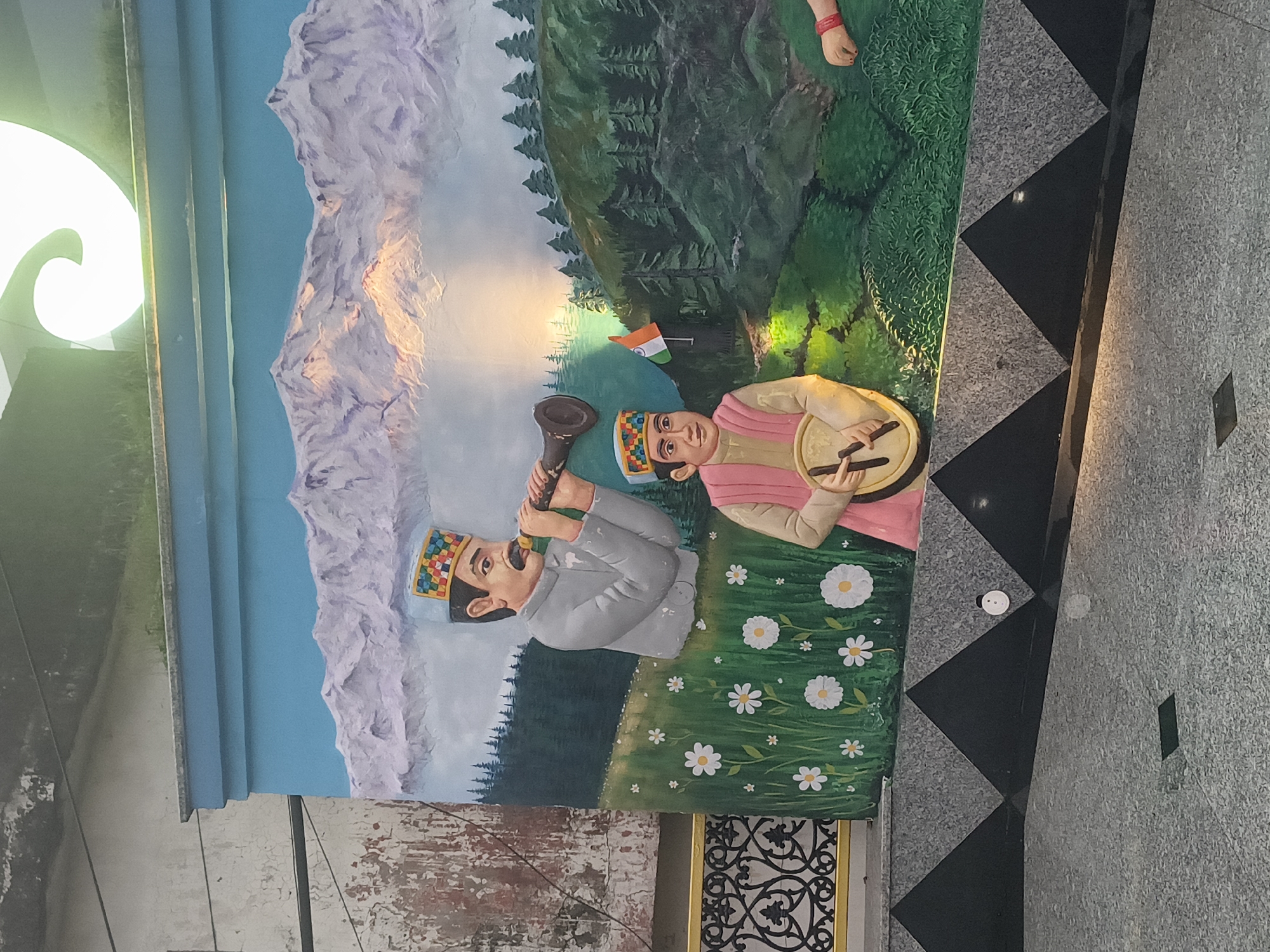 This is a detailed, matter-of-fact description of a bas-relief mural located in Dharamshala, India. The mural depicts a serene mountain landscape with two figures, likely representing local musicians. The background features snow-capped mountains under a soft blue sky, with a setting sun casting warm light on the peaks. The middle ground shows a lush green hillside dotted with pine trees and white flowers.
In the foreground, two male figures are prominently displayed. The older figure, positioned on the left, wears a traditional cap and a grey jacket and is shown playing a wind instrument. To his right, a younger figure, also wearing a traditional cap and a pink vest over a cream-colored tunic, is playing a tambourine. An Indian flag is planted on a small mound between them, adding a patriotic element. The mural is mounted on a textured grey stone base with a decorative black and grey triangular pattern. The overall scene evokes a sense of cultural heritage and natural beauty, typical of the Himalayan region.