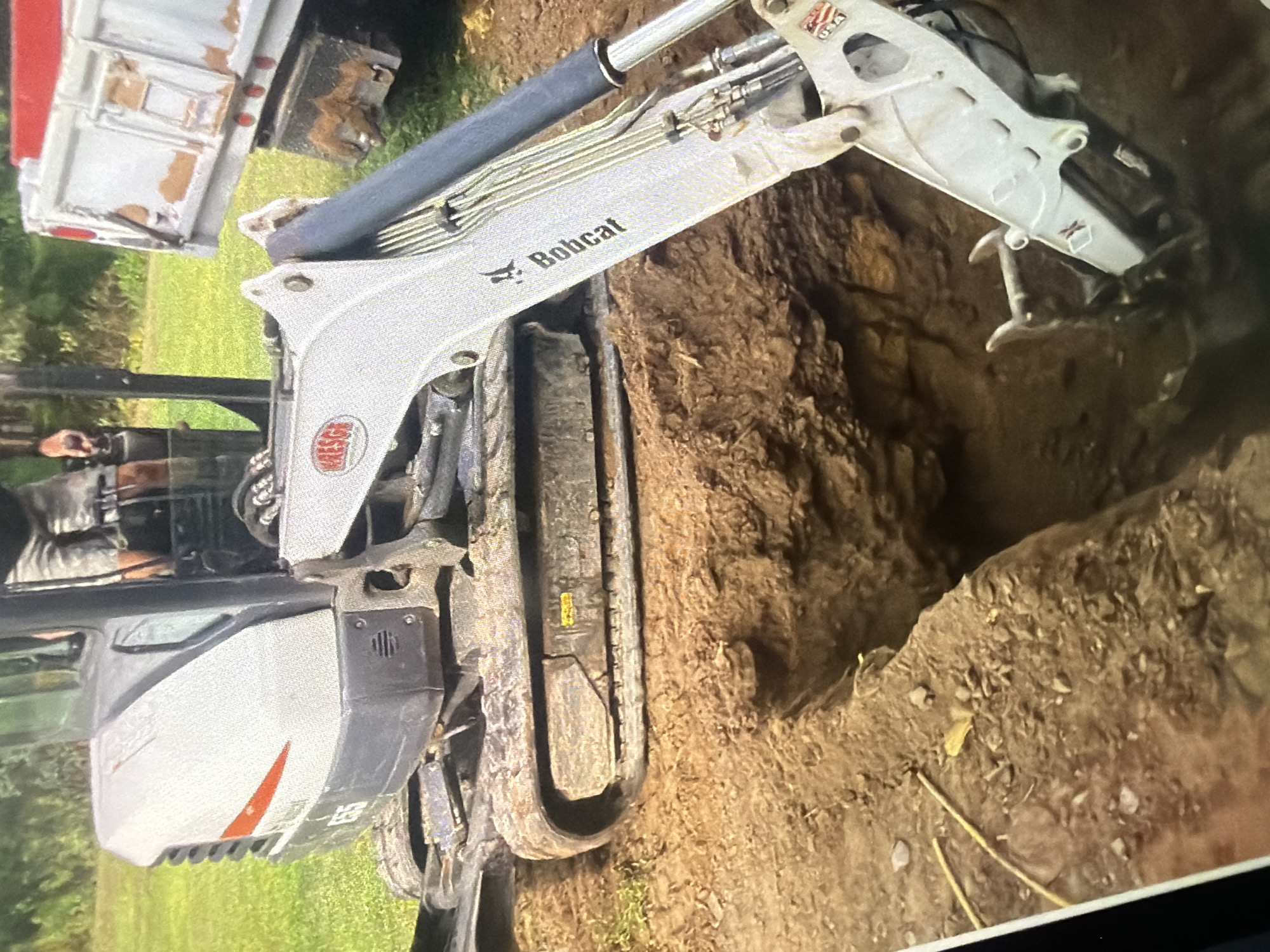 In the city of Unknown, Unknown, an excavator is digging a trench. The excavator is white and has "Bobcat" written on its arm. A person wearing shorts and gloves is operating the excavator. In the background, a dump truck is visible. The ground is dirt and grass.