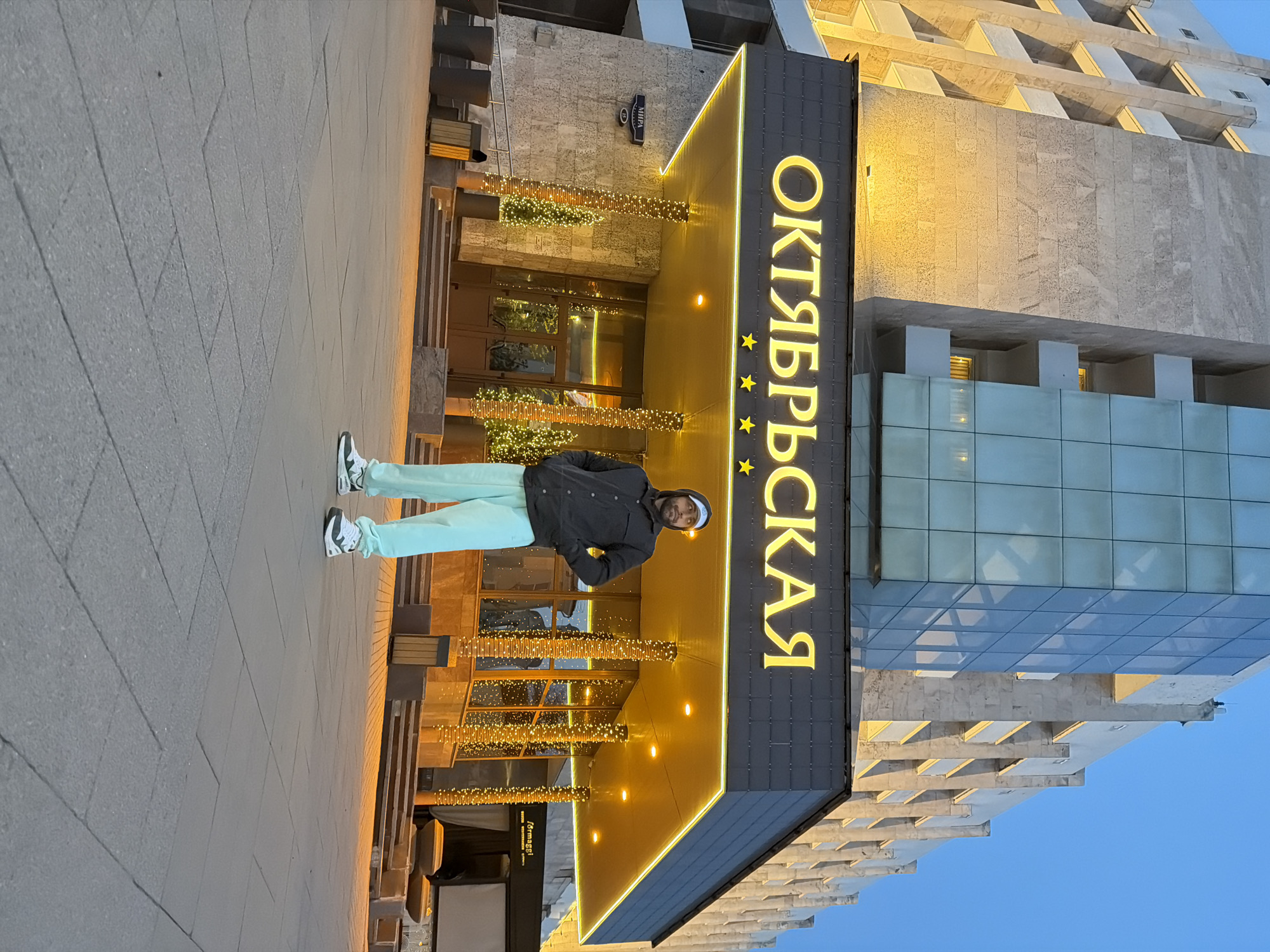 A man is standing in front of the Oktyabrskaya Hotel in Krasnoyarsk, Russia. The hotel's entrance is decorated with lights. The building's architecture is visible in the background. The sky is a pale blue. The man is wearing a dark jacket, light-colored pants, and sneakers.