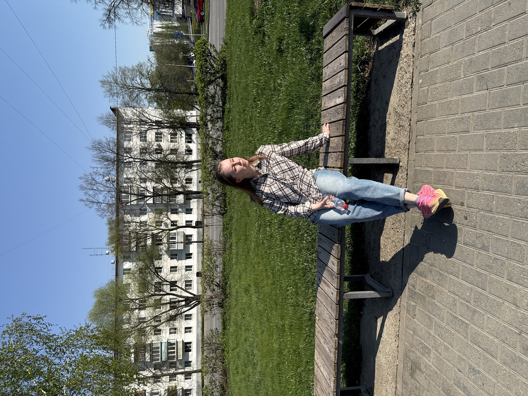 A medium shot captures a woman reclining on a wooden bench outdoors on a sunny day. The woman, wearing a plaid shirt and jeans, holds a plastic water bottle. She is seated on the bench which is adjacent to a paved pathway and a grass lawn, with an apartment building in the background. The setting appears to be in an urban park.