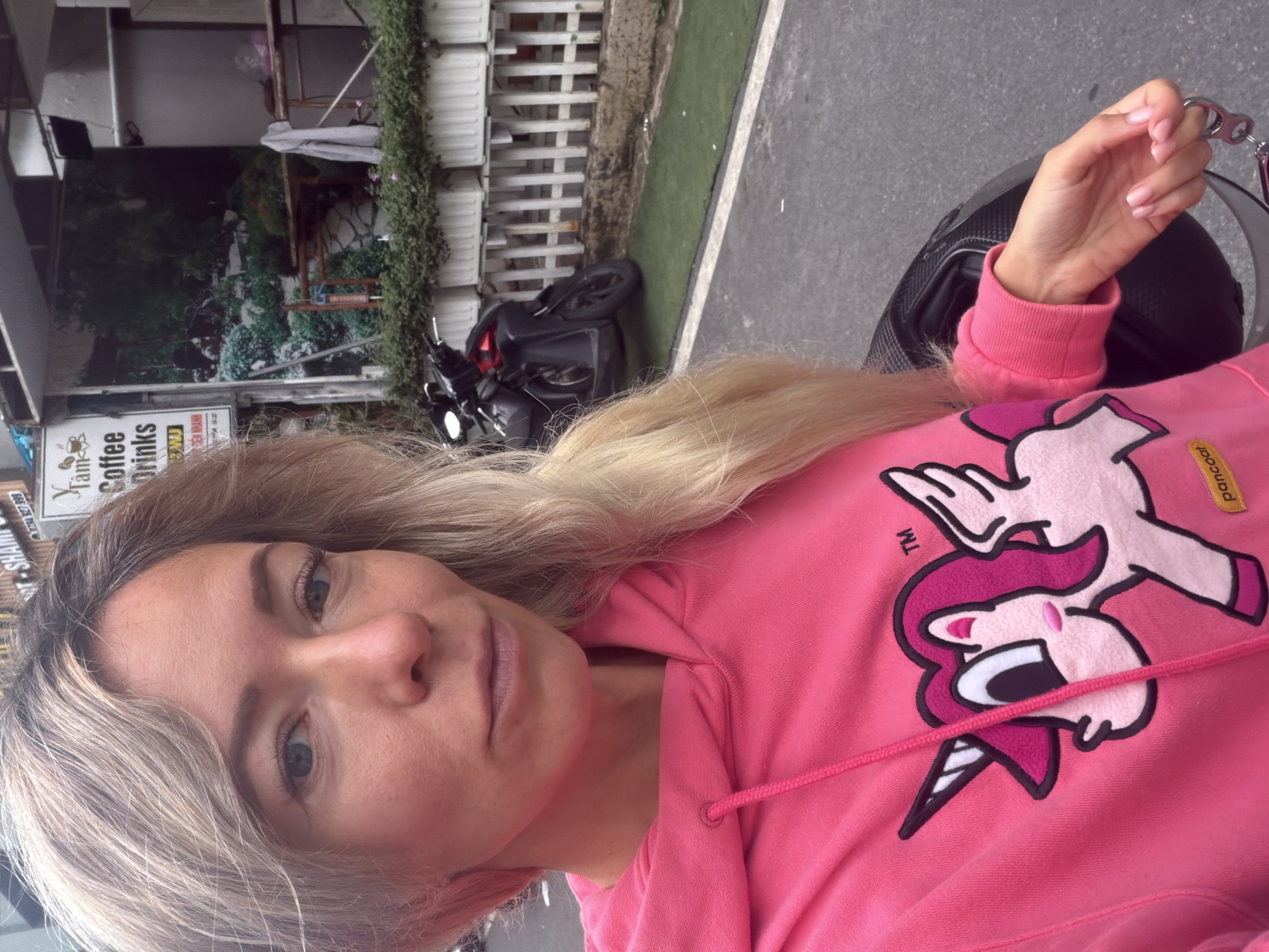A young blonde woman with blue eyes and light skin is taking a selfie in front of a building in Da Lat, Vietnam. She is wearing a bright pink hoodie with a cartoon unicorn design. Her hair is blonde and falls over her shoulders. The background shows a building with a sign that says "Tam Coffee Drinks". There is also a black motorcycle parked in front of the building and some greenery. The woman is holding a keyring in her right hand. The overall impression is a casual outdoor portrait taken on a street in Da Lat.