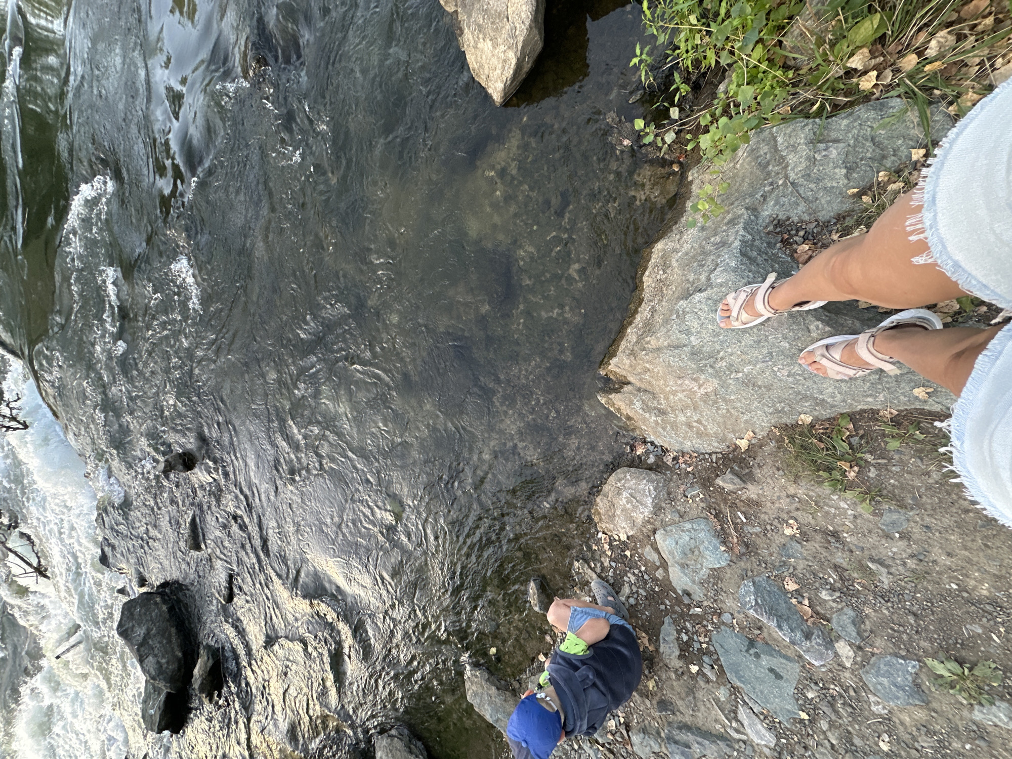 The image depicts a boy sitting on the rocky bank of a river. The boy is wearing a blue cap, a dark hooded jacket, and light-colored shorts. He is looking towards the river. The river is flowing and appears to be shallow in some areas, revealing the rocky riverbed. The bank is covered with stones and some dry leaves. In the foreground, a person's legs are visible, wearing light-colored sandals and denim shorts, indicating they are standing on a large rock overlooking the river. The environment suggests an outdoor natural setting. The location is the city of Energetik, Russia.