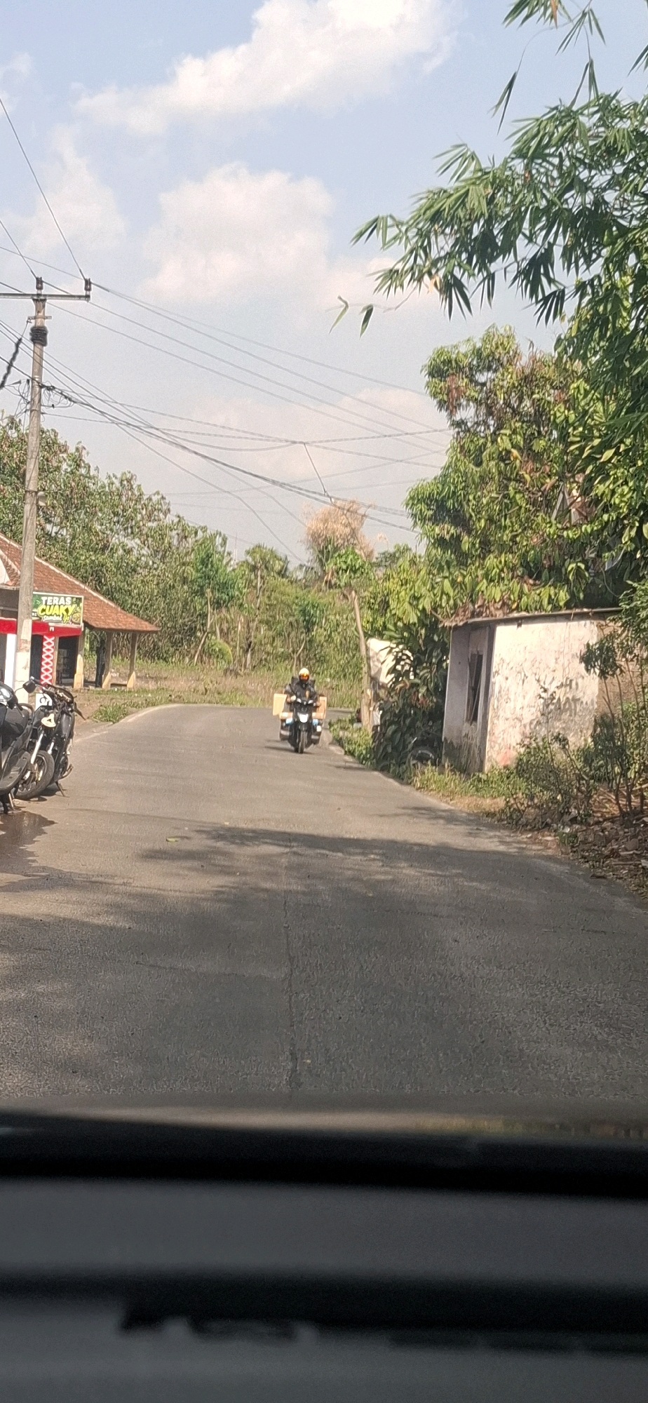 The image captures a street scene in Sirnagalih, Indonesia, on a bright, sunny day with scattered clouds. A paved road, appearing somewhat aged with visible cracks and shadows, curves through a rural or semi-rural area. On the left side of the road, a building with a red roof and white walls is visible, displaying signage with Indonesian text, including "TERAS CUAKY". Parked in front of this building are several motorcycles.
Further down the road, a single individual is riding a motorcycle. The rider is wearing a yellow helmet and a dark jacket, and the motorcycle is equipped with two large, light-colored boxes, suggesting a delivery or transport purpose. The road is flanked by dense green foliage and trees, with a power pole and overhead wires present on the left side. A smaller, dilapidated building with a white, weathered facade and a simple window opening is situated on the right side of the road, partially obscured by vegetation. The overall impression is that of a typical Indonesian roadside environment.