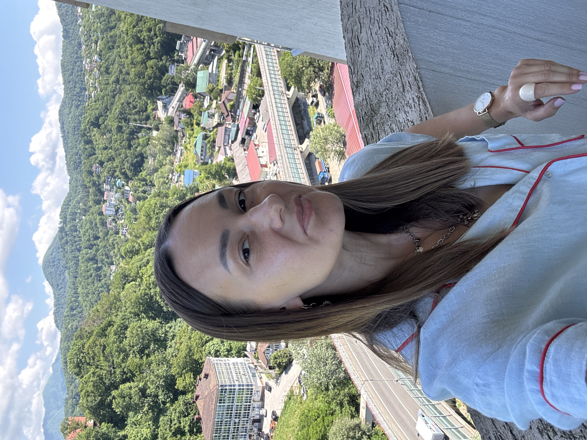 A woman takes a selfie on a balcony overlooking a city nestled among lush green mountains under a blue sky with scattered white clouds. She has long brown hair, light skin, and is wearing a light blue button-down shirt with red trim. She is also wearing a watch on her left wrist and a large ring on her right index finger. In the background, numerous buildings with red and green roofs are visible, along with a highway and an elevated pedestrian bridge. The architecture of the buildings and the mountainous terrain suggest the location is Sochi, Russia, a city known for its Olympic facilities and proximity to the Caucasus Mountains. The scene captures a moment of leisure and sightseeing in a picturesque, urban-mountain landscape.