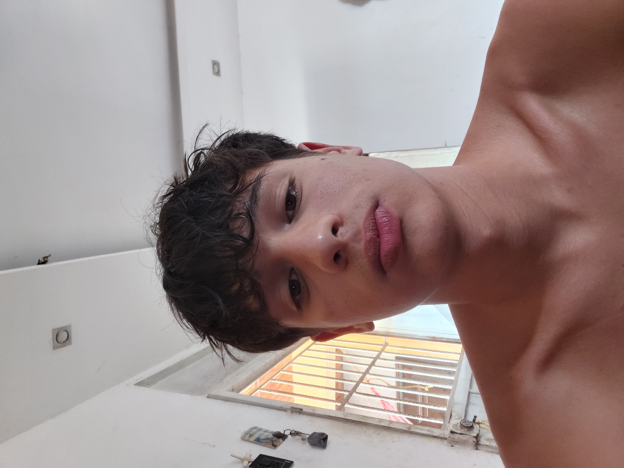 A close-up, head-and-shoulders selfie of a young male with dark, messy hair and light brown eyes is presented. He has a fair complexion with some minor blemishes and is looking directly at the camera with a neutral expression. His lips are slightly parted. He is shirtless, with his shoulders and upper chest visible.
The background suggests an indoor setting with a white wall and ceiling. A square light fixture is visible on the ceiling. To the left of the subject, a window with a metal grate is partially visible, with some indeterminate objects hanging on the wall next to it. The overall lighting appears to be natural, possibly from the window, casting a warm tone on the subject's skin. The setting is consistent with a residential interior in a city like Santa Ana, Venezuela.