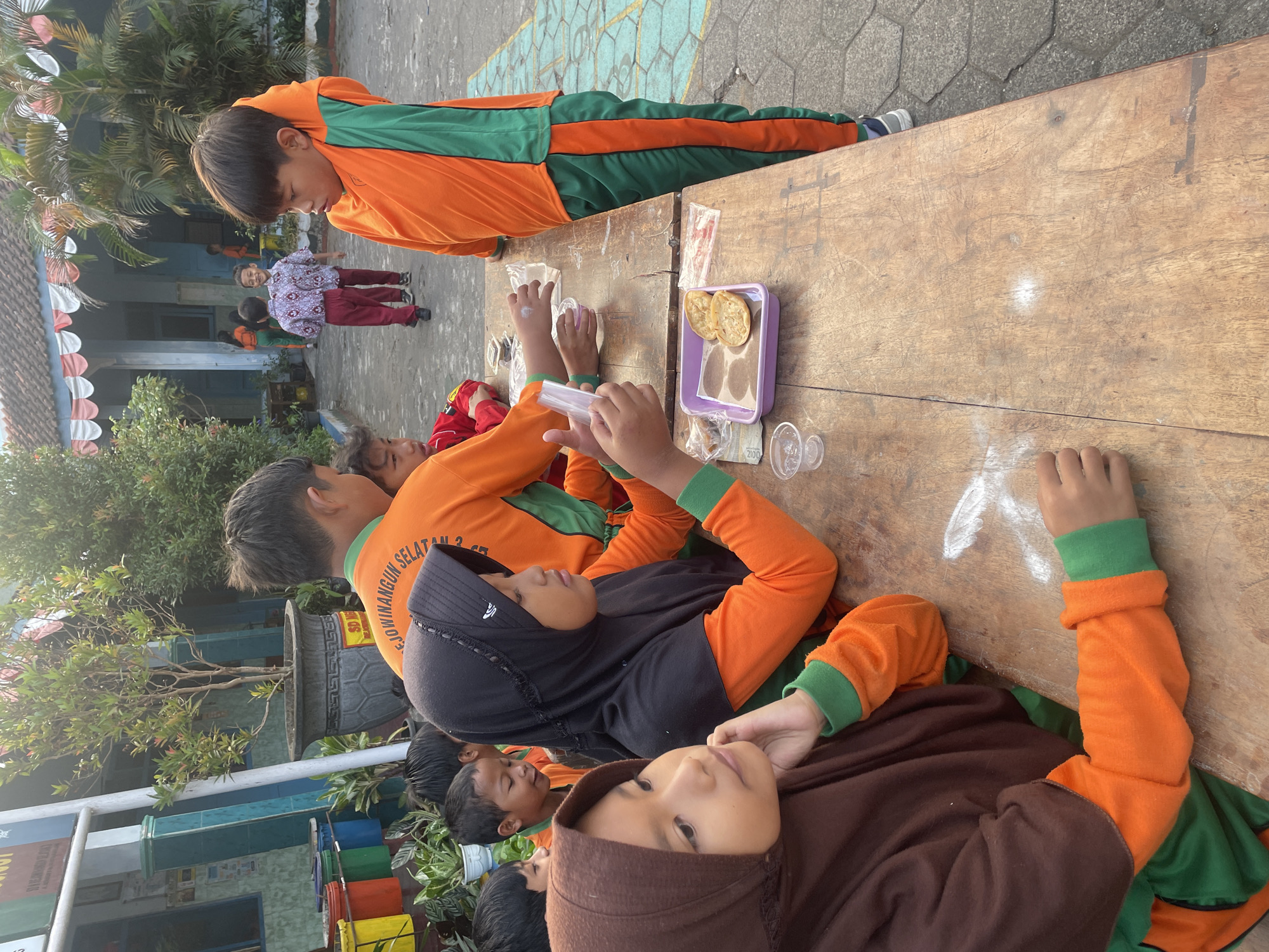 The image depicts a group of children, likely students, gathered around a wooden table outdoors. Most of the children are wearing matching orange and green uniforms. Some are engaged in activities on the table, while others are looking around. The scene appears to be in a schoolyard in Magelang, Indonesia, given the architectural style of the building in the background, the type of foliage, and the children's attire, which is common for Indonesian school children.
In the foreground, two girls are seated at the table. The girl on the left, wearing a brown hijab, looks to her left with a thoughtful expression, her hand resting on her chin. Beside her, a girl in a black hijab and orange uniform is looking down at the table, her hand near a small plastic container and some food items. Other children in similar uniforms are visible around the table, some appearing to interact with each other or the items on the table.
On the table, there are several objects including small plastic cups, a purple tray with what appear to be fried snacks, and some small packets. The table itself is weathered and appears to be made of wood.
The background shows a school building with a tiled roof and columns, and lush green trees and plants. The ground is paved with stone. The overall atmosphere suggests a daytime activity or event taking place at the school.