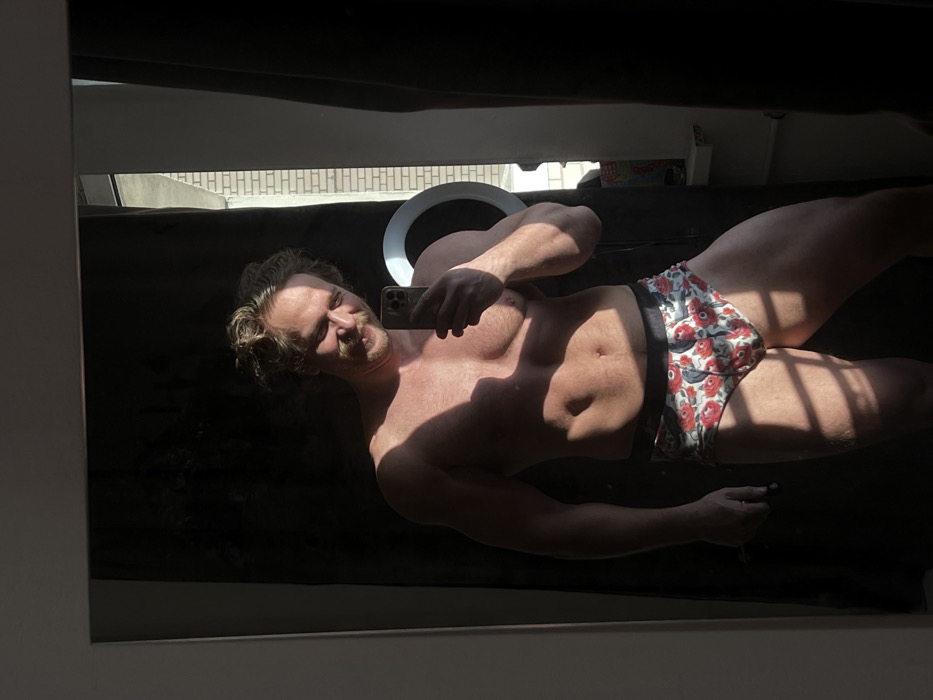 An adult male, light-skinned with wavy light brown hair and a mustache, is captured in a mirror selfie. He is smiling with squinted eyes. The subject is bare-chested and wears white briefs adorned with a red and green floral pattern and a dark waistband. He holds a dark-colored smartphone, consistent with an iPhone model, in his right hand, angled to capture his reflection. Strong natural light, originating from the right side of the frame, illuminates the scene, casting distinct shadows across his torso, legs, and the immediate floor area. Visible in the reflection behind his right shoulder is a white ring light. The background behind the subject is predominantly dark, appearing to be a dark curtain or fabric. To the right, a window reveals a portion of an exterior light-colored brick building, a white window sill, and a vertical white pipe. The scene is located in the city of Saint Petersburg, Russia.