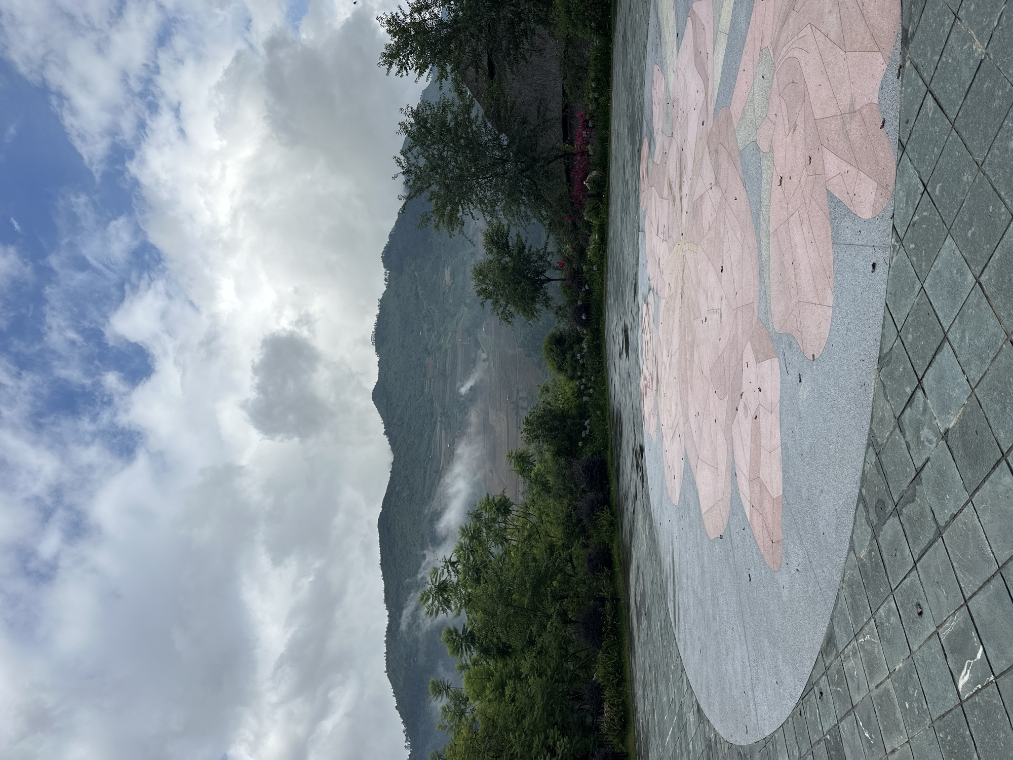 The scene captures an outdoor setting in Mu Cang Chai, Vietnam, under a mostly overcast sky with hints of blue. In the foreground, a paved plaza comprises dark grey-green square tiles forming an outer perimeter and a central, lighter grey circular section adorned with a large, stylized pink and light grey floral or leaf mosaic pattern. Scattered dry leaves lie on the damp paved surfaces. A dense band of green trees and shrubs separates the plaza from the mountainous background. The prominent mountains exhibit extensive terracing on their slopes, characteristic of rice paddies, partially enveloped by low-lying mist and clouds. On the far right, a structure with a distinctive thatched roof is visible behind the trees. No individuals are present in the image.