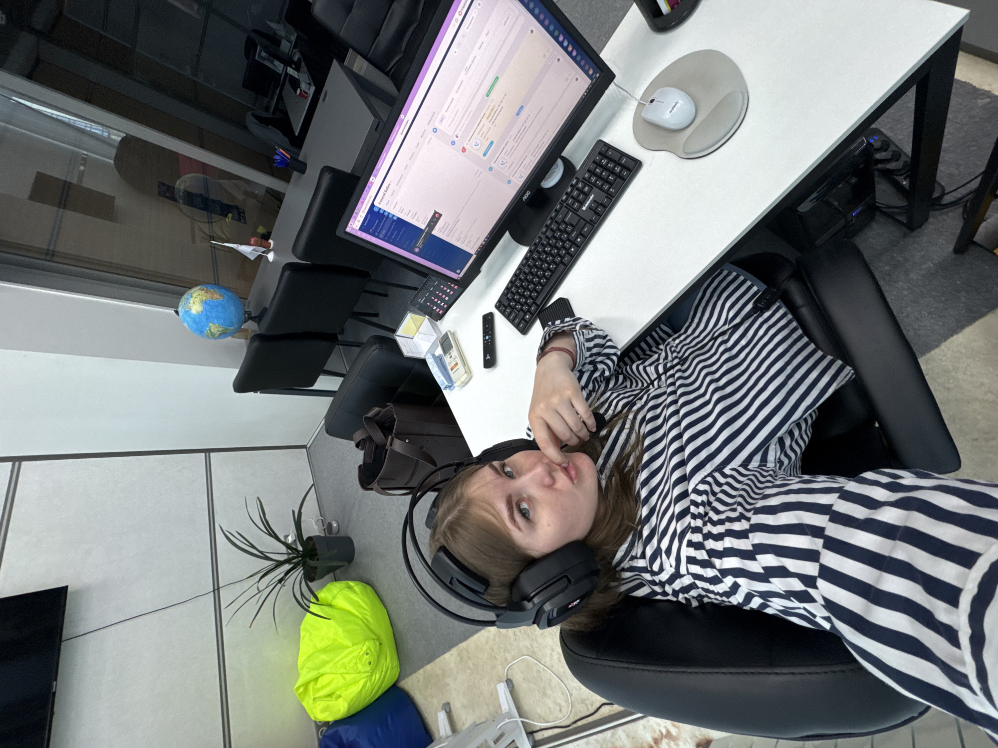 The image shows a person sitting at a desk in what appears to be an office setting. The location is Perm, Russia.
Subject:
- A person wearing a black and white striped shirt and headphones.
- Desk and chair.
Objects:
- Computer monitor displaying a software interface.
- Keyboard, mouse, and mousepad on the desk.
- Globe, office supplies, and remote controls on the desk.
- Potted plant, beanbag chairs, and a TV on the floor.
Environment:
- Office setting with tiled flooring and a window.
- Desk with a white top and black frame.
- Black office chair.
Actions:
- The person is sitting in the chair and facing the camera.
Interaction:
- The person is interacting with the office environment.