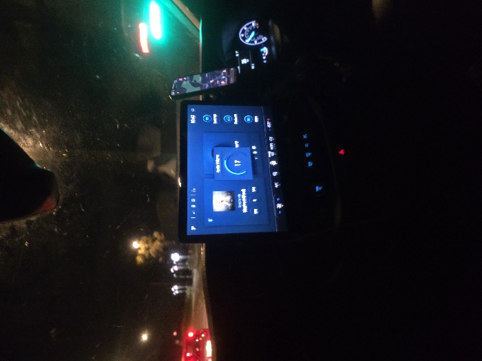 The image displays the interior of a vehicle at night, looking forward through a smudged windshield, situated in Woking, United Kingdom. A central digital infotainment screen, emitting a blue light, dominates the dashboard, showing a music player interface with an album cover, a numerical volume indicator of '17', and a digital clock displaying '10:52'. Below this screen, multiple blue-illuminated control buttons are visible, including a red triangular hazard light button. To the right of the infotainment display, a smartphone in a camouflage-patterned case is vertically mounted. A segment of the vehicle's instrument cluster, featuring a blue-lit dial with an active needle, is also visible on the right side of the frame. Outside the vehicle, the red taillights of another car are visible on the left. The road ahead is illuminated by streetlights, with a yellow-lit tree further in the distance. A prominent bright green traffic light is visible in the upper right portion of the external view. No individuals are depicted within the scene.