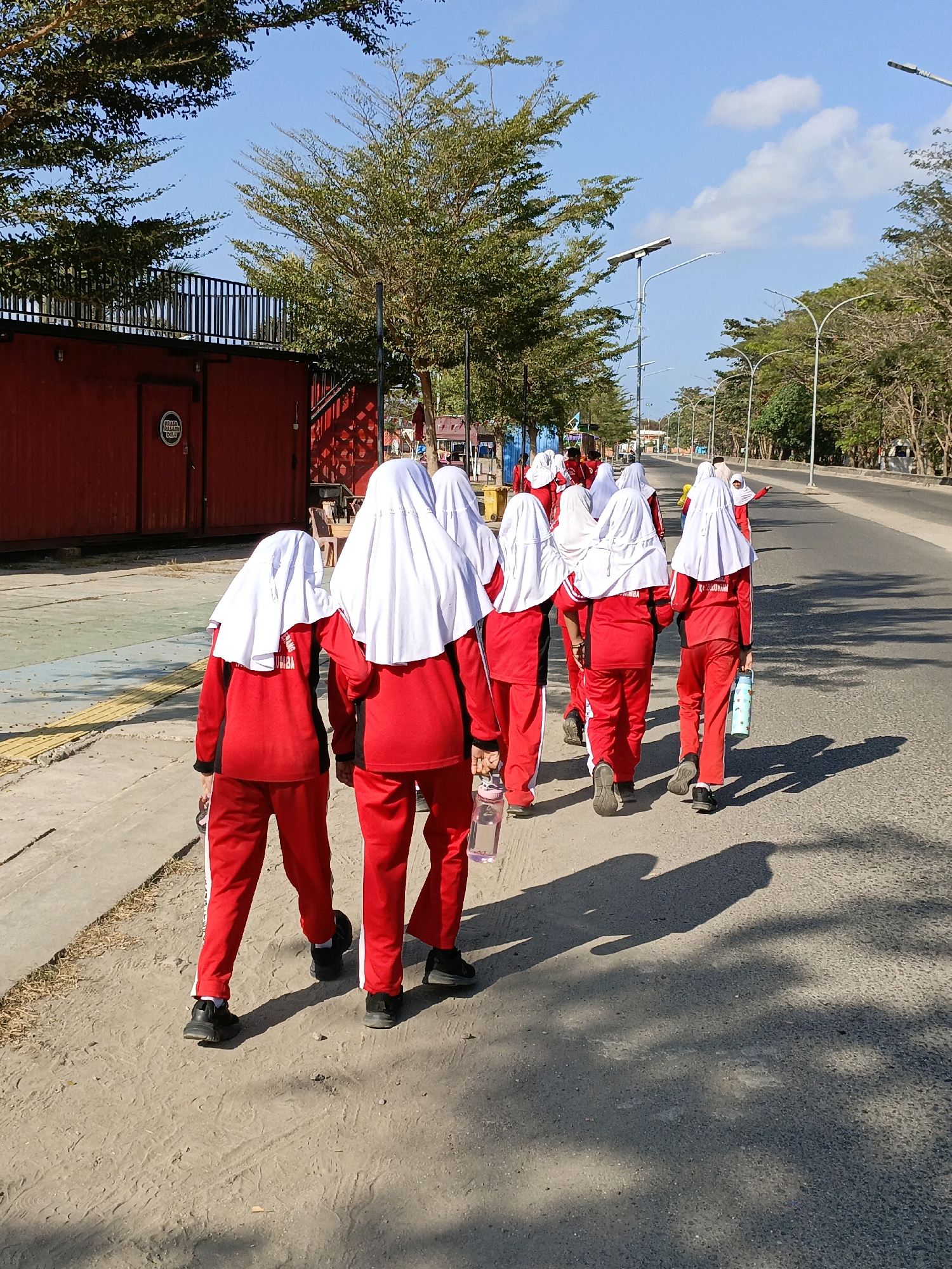 A group of eight children, all wearing matching red tracksuits and white headscarves, walk away from the camera on a paved road in Bulukumba, Indonesia. The children are carrying water bottles. The road is lined with trees on the right side, and on the left is a red building with a black railing on top, suggesting it might be a container-style building used for commerce or shelter. The sky is clear and blue with a few scattered clouds. The scene appears to be outdoors during daylight.