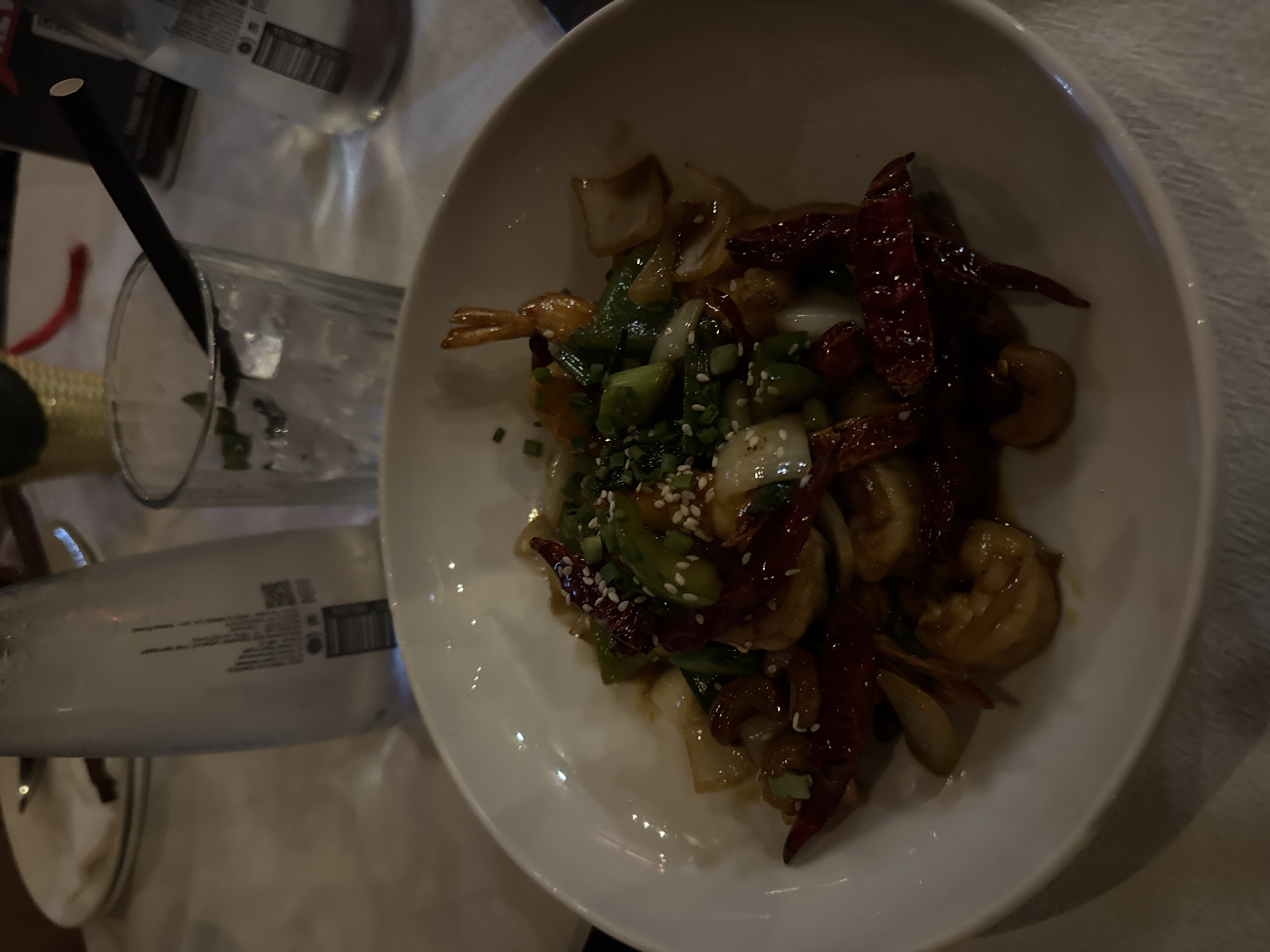 A white, shallow bowl containing a stir-fry dish is centered in the foreground, resting on a white tablecloth. The dish is composed of shrimp, dried red chili peppers, green bell peppers, and onions, all coated in a glossy sauce and sprinkled with sesame seeds and chopped green onions. To the left of the bowl, partially visible, is a clear glass filled with ice and a black straw, positioned next to a clear glass bottle of water. In the background, a gold woven basket and a black menu with red lettering are discernible. The overall lighting suggests an indoor setting, possibly a restaurant in Kuta, Indonesia, given the context.
