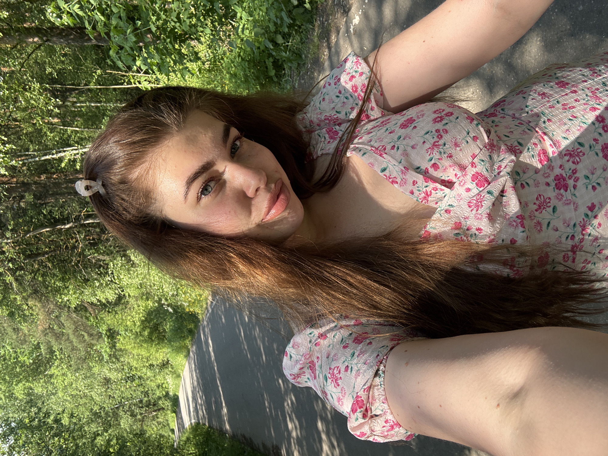 In the city of Shavlyagino, Russia, a young woman with long brown hair, adorned with a translucent clip, poses for a selfie on a paved path. She is wearing a light-colored dress with a pink floral pattern. The path winds through a lush forest filled with green trees, casting dappled shadows on the ground. Sunlight filters through the leaves, illuminating the scene and highlighting the woman's features. Her left arm is extended towards the camera, holding the device to capture the image. The background consists of a dense, verdant forest with sunlight illuminating the trees and foliage.