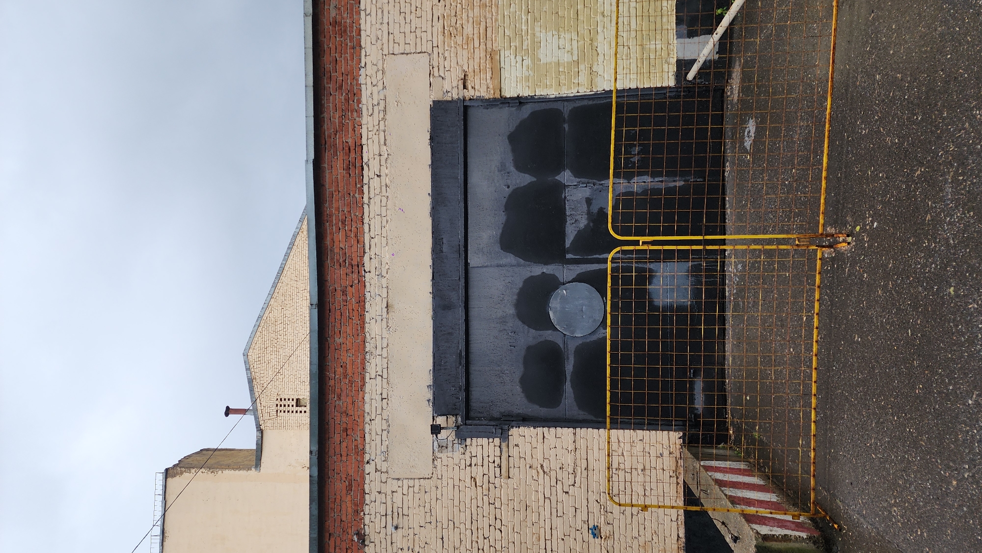 The image depicts a building in Moscow, Russia. The building's exterior walls consist of beige and red bricks, with some sections showing signs of paint peeling or aging. A large, black, metal door is centered on the building's facade. The door is wet, suggesting recent rain. In front of the door, a yellow metal barrier is placed. The barrier is on an asphalt surface, which also appears wet. The sky is overcast, indicating cloudy weather. The surrounding structures suggest an urban environment.