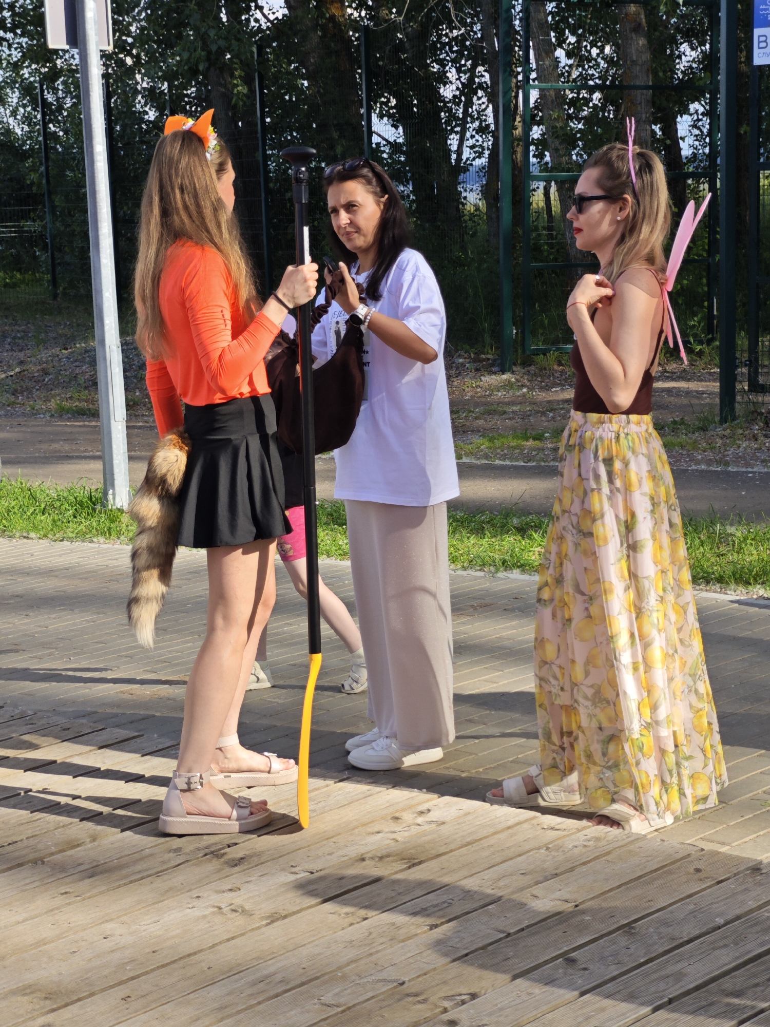 Three people are standing on a wooden boardwalk on a sunny day. On the left, a young woman with long brown hair wears a bright orange long-sleeved shirt, a short black skirt, and a fluffy, bushy tail attached to her lower back. She also has orange fox ears on a headband. She is holding a long, dark staff with a yellow tip. In the center, a woman with dark hair wears sunglasses on her head and a white t-shirt with a graphic design. She is holding a smartphone and a dark brown handbag. She is wearing loose-fitting beige pants. On the right, a woman with blonde hair pulled back in a ponytail wears a brown sleeveless top and a long, flowing skirt with a yellow lemon print. She has pink fairy wings attached to her back and is wearing sunglasses. The background shows trees and a green fence. The scene appears to be outdoors, possibly at a park or an event. The lighting suggests it is daytime, and the weather is clear and sunny.