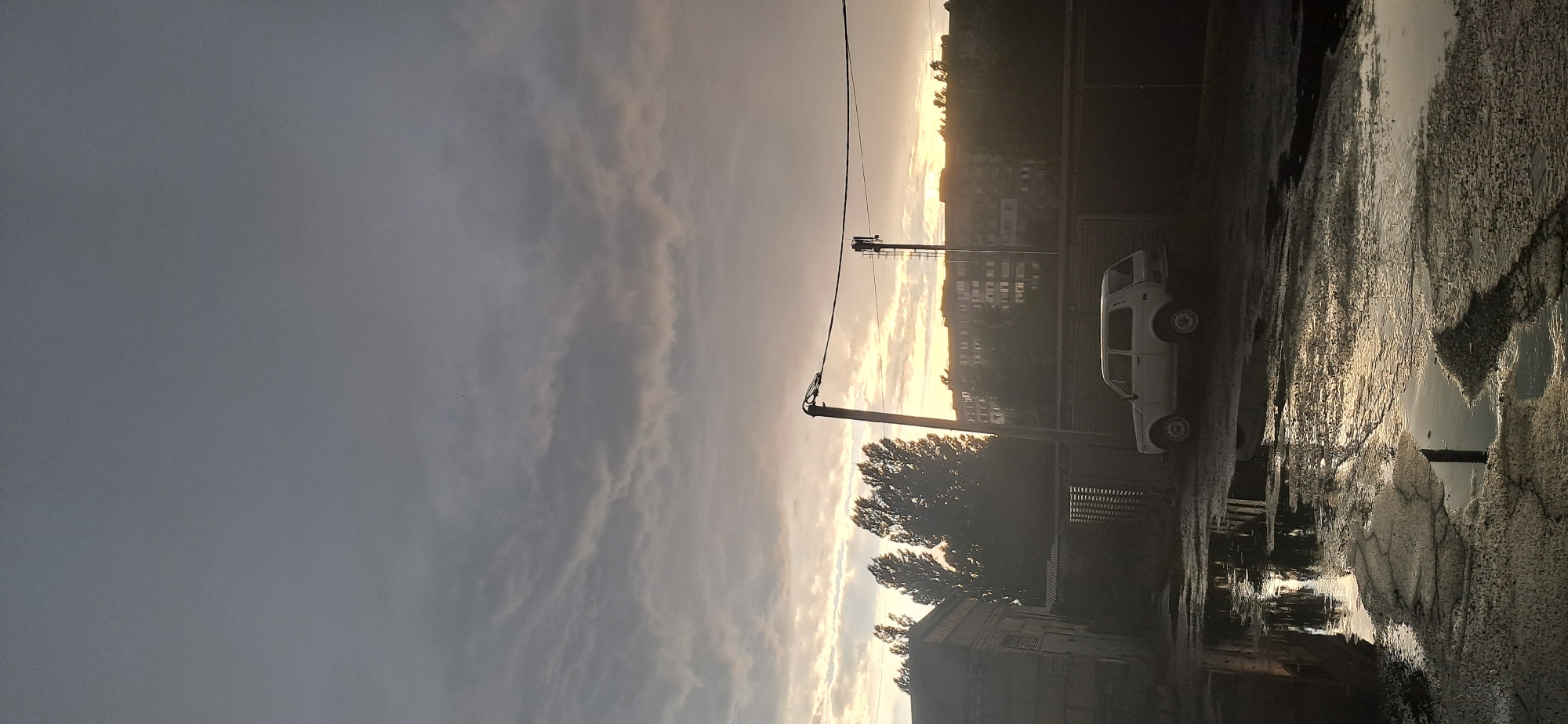 This is a vertical, medium-shot photograph taken outdoors in Horishni Plavni, Ukraine. The scene depicts a street or courtyard area under a cloudy sky with the sun setting or rising, casting a warm glow. In the foreground, the ground is wet and reflective, with several large puddles of water indicating recent rain or a leak. A white SUV is parked on the right side of the frame, facing towards the left. Behind the car, there is a dark fence and further back, trees and buildings are visible. A street light pole stands in the middle ground, with power lines stretching across the scene. To the left, a brick building is partially visible. The sky is filled with dramatic, textured clouds that are lighter at the horizon, suggesting the sun is low in the sky. There are no people present in the image. The overall impression is one of a quiet, perhaps somewhat neglected, urban environment during twilight.