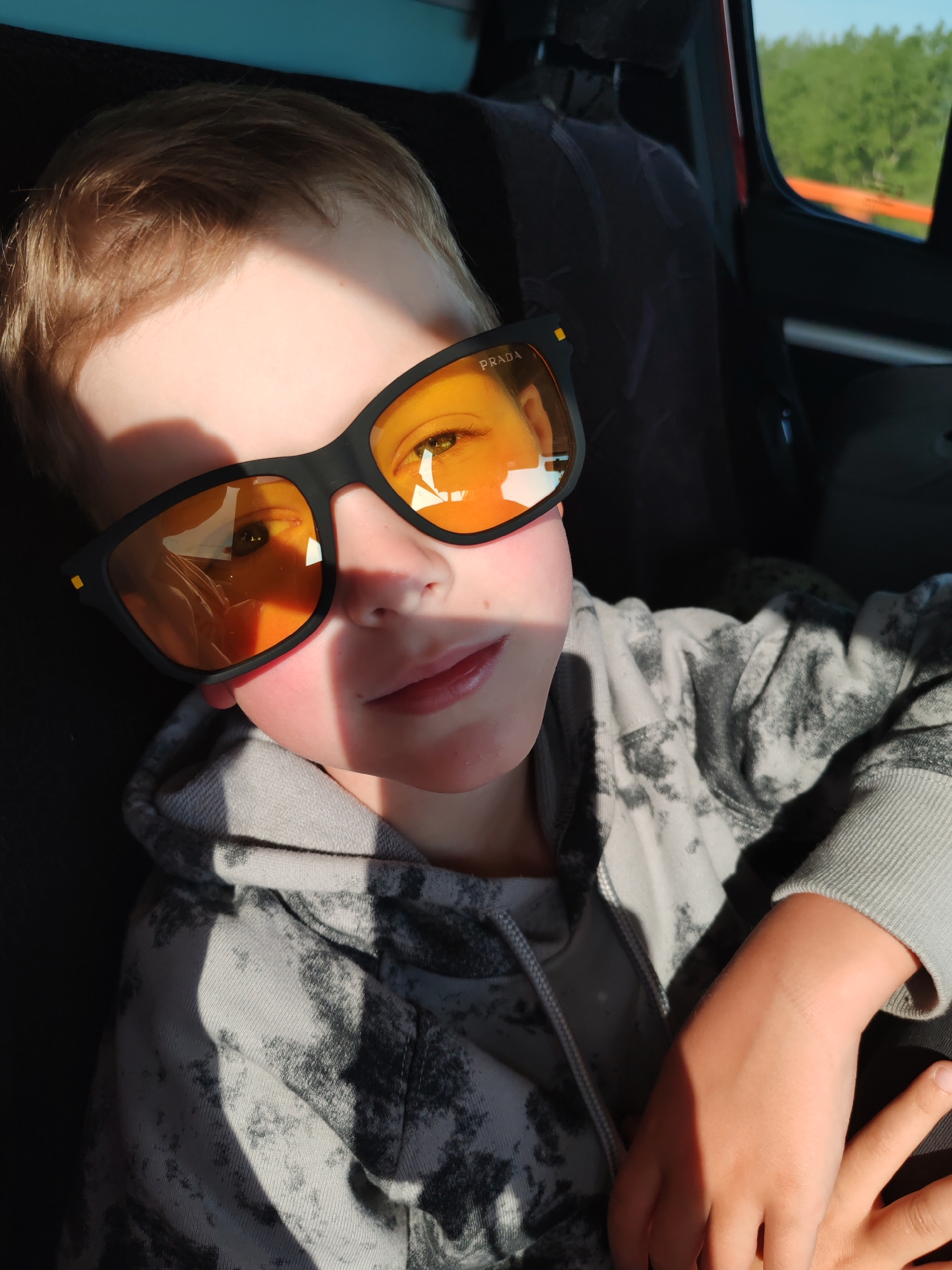 A young boy is sitting inside a vehicle, likely a car, wearing a grey tie-dye hoodie and black Prada sunglasses with orange lenses. Sunlight casts a shadow across the upper portion of his face. The car's interior is visible behind him, including the seat and headrest. In the reflection of the sunglasses, it appears he is traveling through a rural environment. The picture was taken in Diurtiuli, Russia.