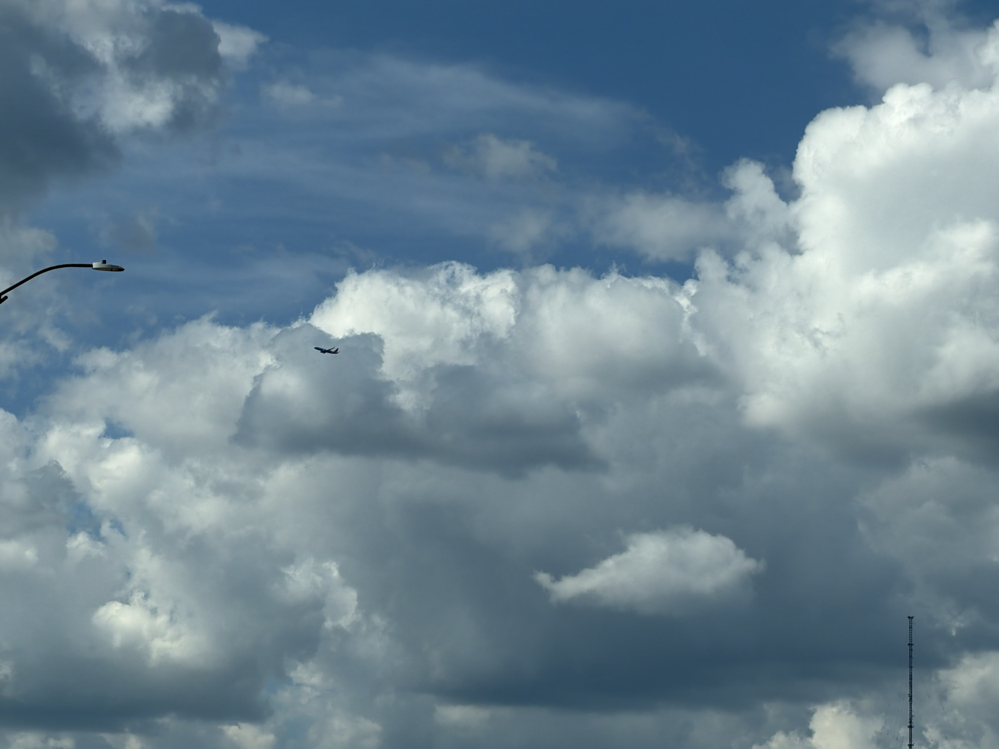 The image shows a bright blue sky with a variety of clouds. There is a single airplane visible in the distance, flying horizontally across the frame. In the lower left corner, a street lamp post curves into the frame. In the lower right corner, a tall, slender radio tower is visible against the clouds. The scene appears to be an outdoor daytime shot, possibly during the day. No specific location in Pittsburgh can be determined from the image.