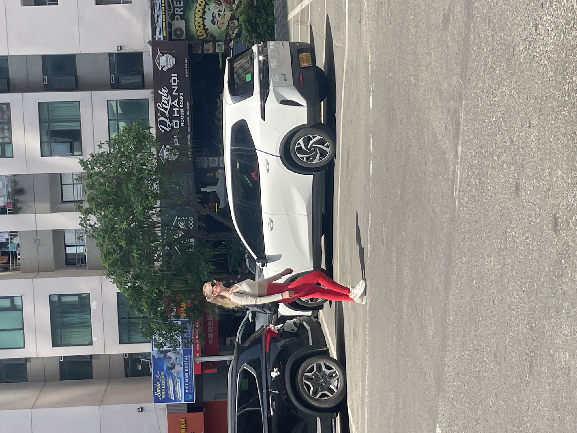 A blonde woman in a white crop top and red leggings walks in a parking lot. She is wearing sunglasses and white sneakers. To her left, a black SUV is parked. To her right, a white SUV is parked. In the background, there are multi-story buildings with windows. A sign for "D'LINH PHO HA NOI NOODLE SOUPS" is visible. The location is Nha Trang, Vietnam.