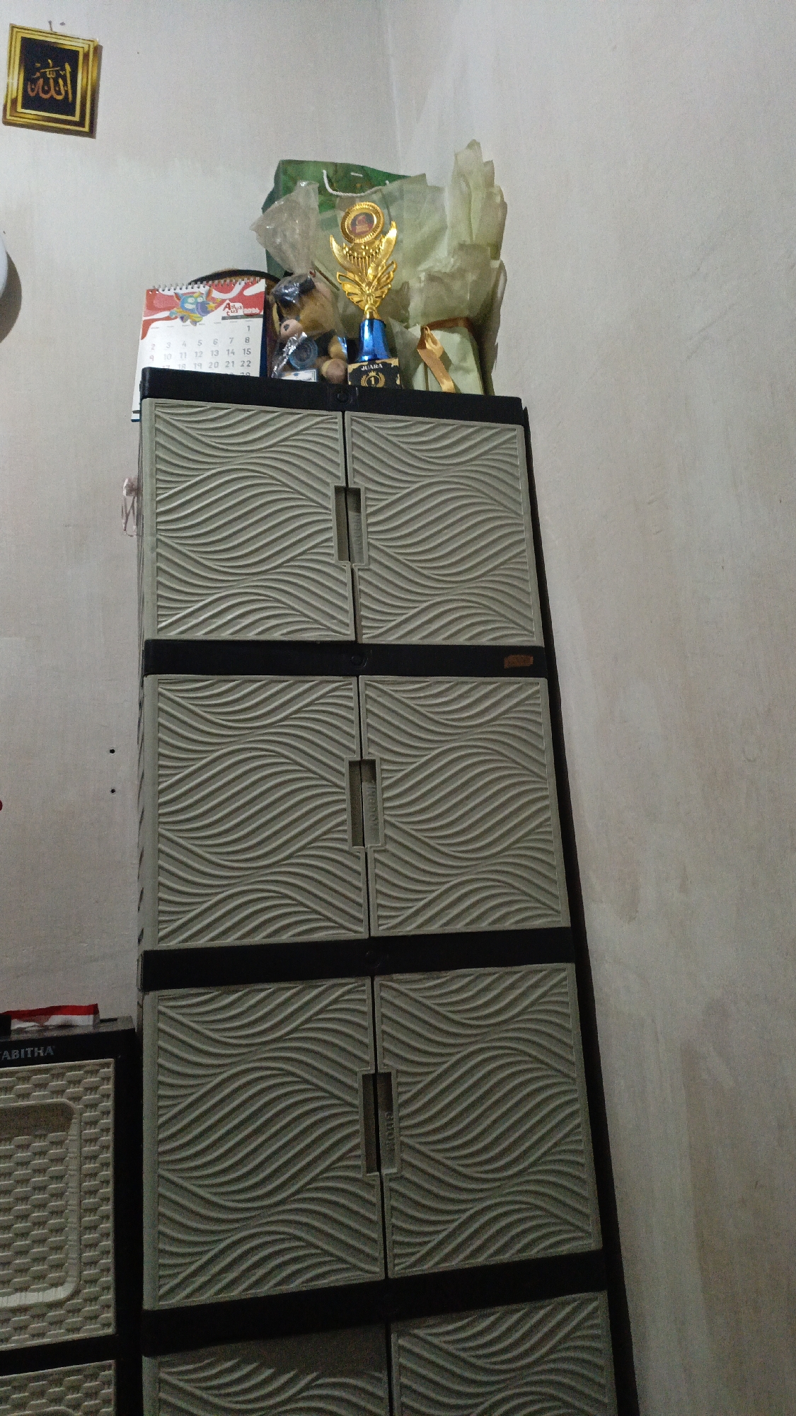 A trophy, a teddy bear, a bouquet, and a calendar are displayed on top of a plastic cabinet. The cabinet is tall and has four doors, each with a wavy pattern. To the left of the cabinet is another plastic storage unit with a basket-like texture. On the wall above the cabinet, a framed Arabic calligraphy art piece is hung. The scene appears to be indoors, in a room with light-colored walls. The overall setting is a domestic environment, possibly a home in Pejala, Indonesia.