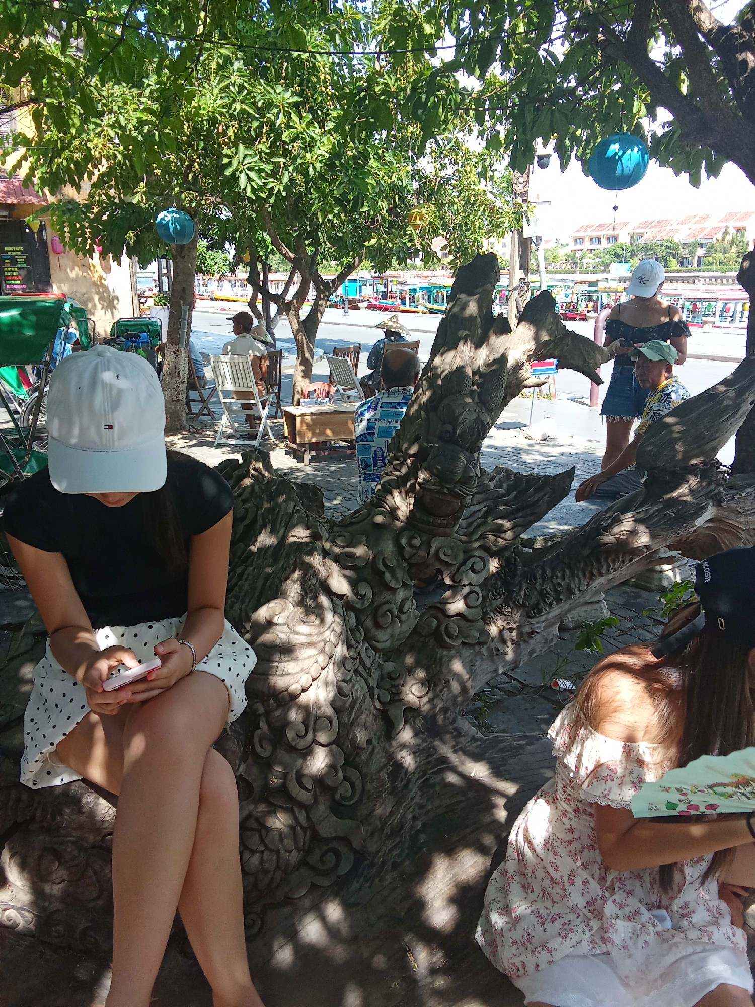 In Hội An Ward, Vietnam, a woman wearing a white baseball cap and a black off-the-shoulder top sits with her legs crossed, looking down at a pink smartphone. She wears a white skirt with black polka dots. In the foreground, a large, intricately carved wooden sculpture is visible. Behind her, other people are seated at tables and chairs, some wearing hats. The background shows a street scene with buildings, trees, and boats on a waterway. Blue lanterns hang from tree branches. The scene appears to be an outdoor seating area, possibly a cafe or restaurant, with a focus on cultural art and leisure.