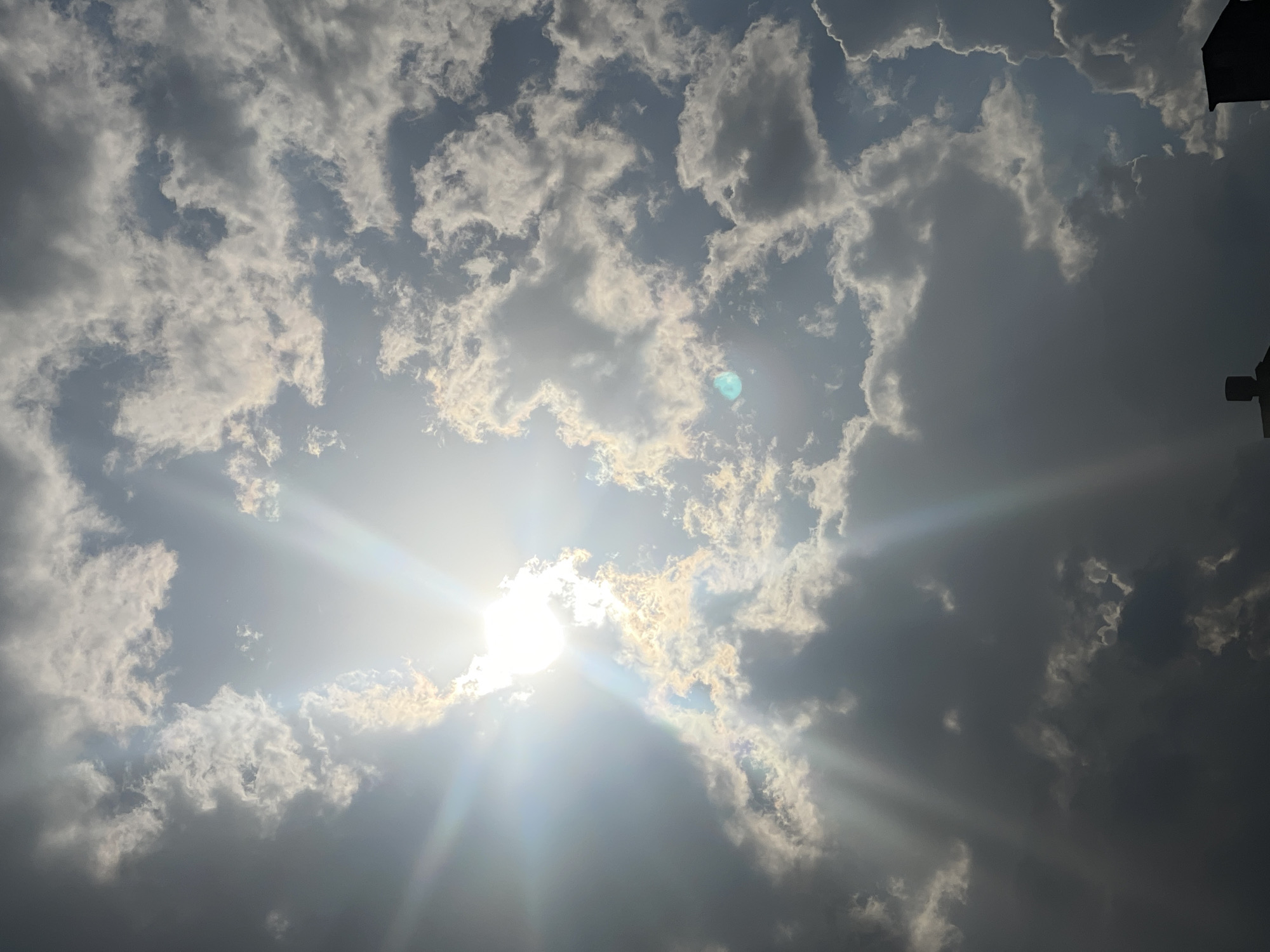 This image captures a dramatic sky over the city of Varna, Bulgaria. The sun, partially obscured by clouds, shines brightly in the upper left quadrant, casting luminous rays that pierce through the textured cloud formations. The clouds themselves are a mix of dark, heavy masses and lighter, illuminated wisps, creating a dynamic contrast. The lower portion of the image shows the silhouetted rooftops of buildings, suggesting an urban setting. The overall impression is one of a powerful and active sky.