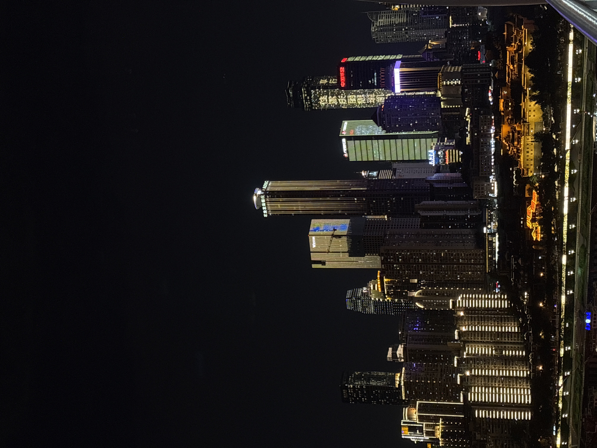 The image depicts a nighttime cityscape in Nan'an District, Chongqing, China, focusing on a cluster of illuminated skyscrapers and other buildings. The dark sky serves as a backdrop for the vibrant artificial lighting emanating from the structures. Several prominent buildings are visible, including a tall skyscraper with a "Westin" sign and a building with vertical golden lights. Other buildings feature different patterns and colors of illumination, such as green, blue, and purple lights. In the foreground, a river or waterway is visible, reflecting the lights from the city. Along the riverbanks, there are trees and lower-rise buildings, some of which are illuminated with warm yellow lights, creating a sense of urban density and activity. The overall scene conveys a modern and dynamic urban environment at night, with a strong emphasis on architectural lighting.